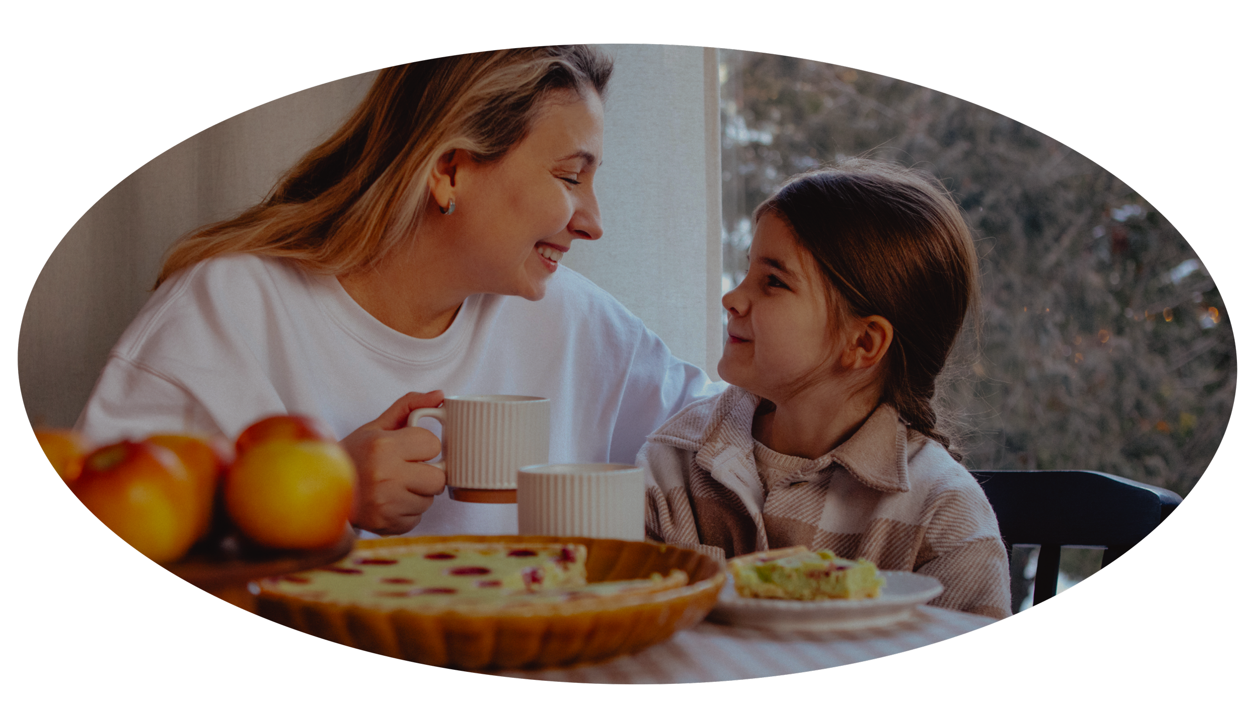 A woman and a young girl smiling at each other at a dining table with apples and a pie.