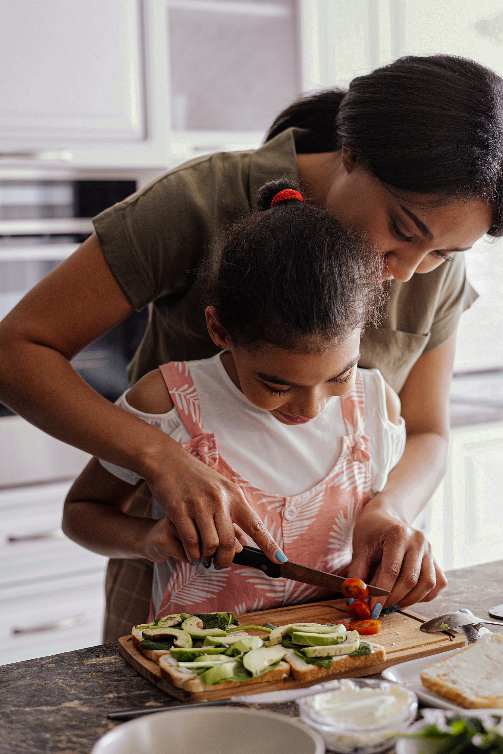 A woman and a young girl preparing a sandwich in a kitchen. The woman is guiding the girl as they cut vegetables on a wooden cutting board. There are bowls of ingredients and slices of bread on the counter.