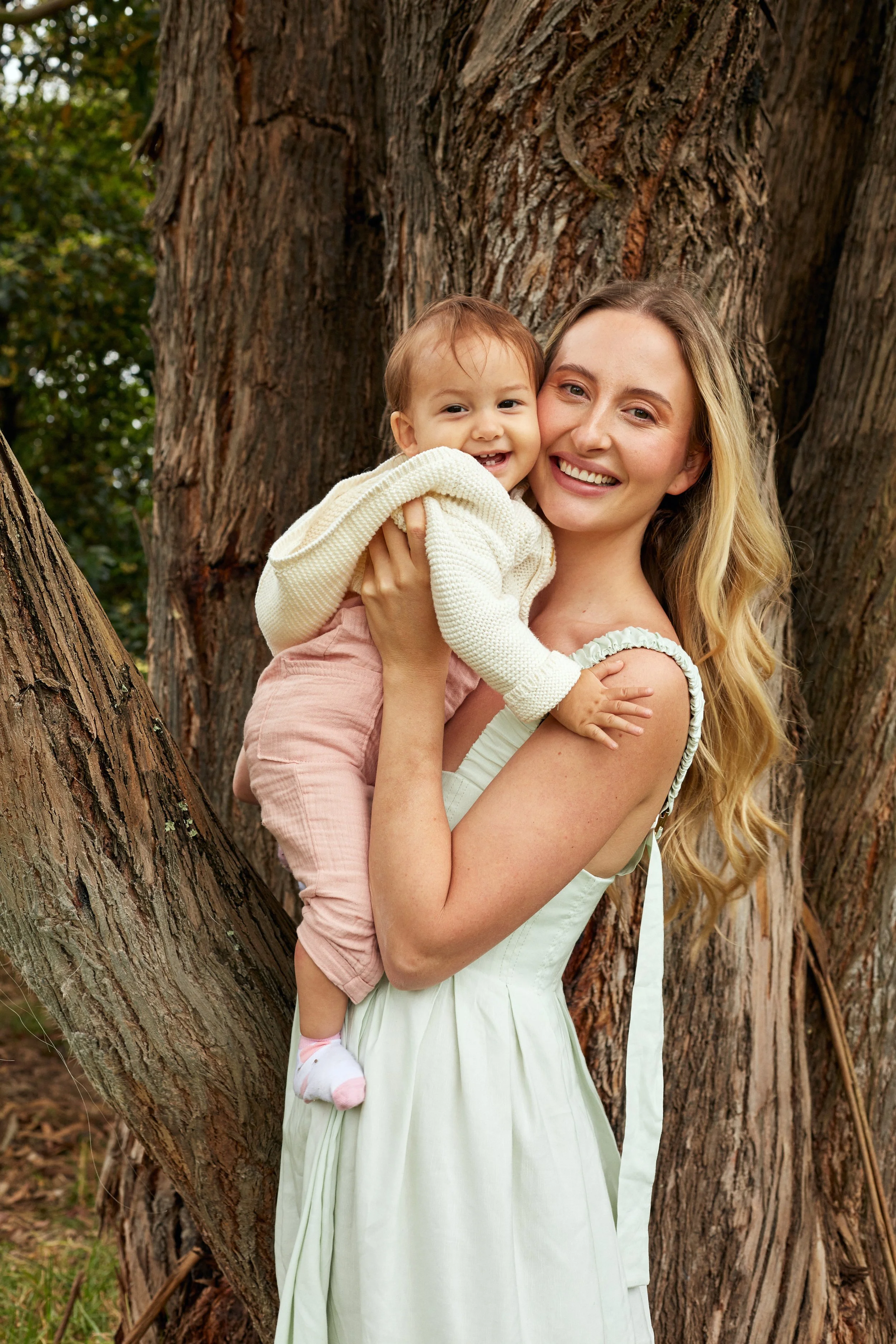 A woman with long blonde hair holding a smiling toddler girl with short light brown hair and light skin in front of a large tree with textured bark, outdoors during daytime.