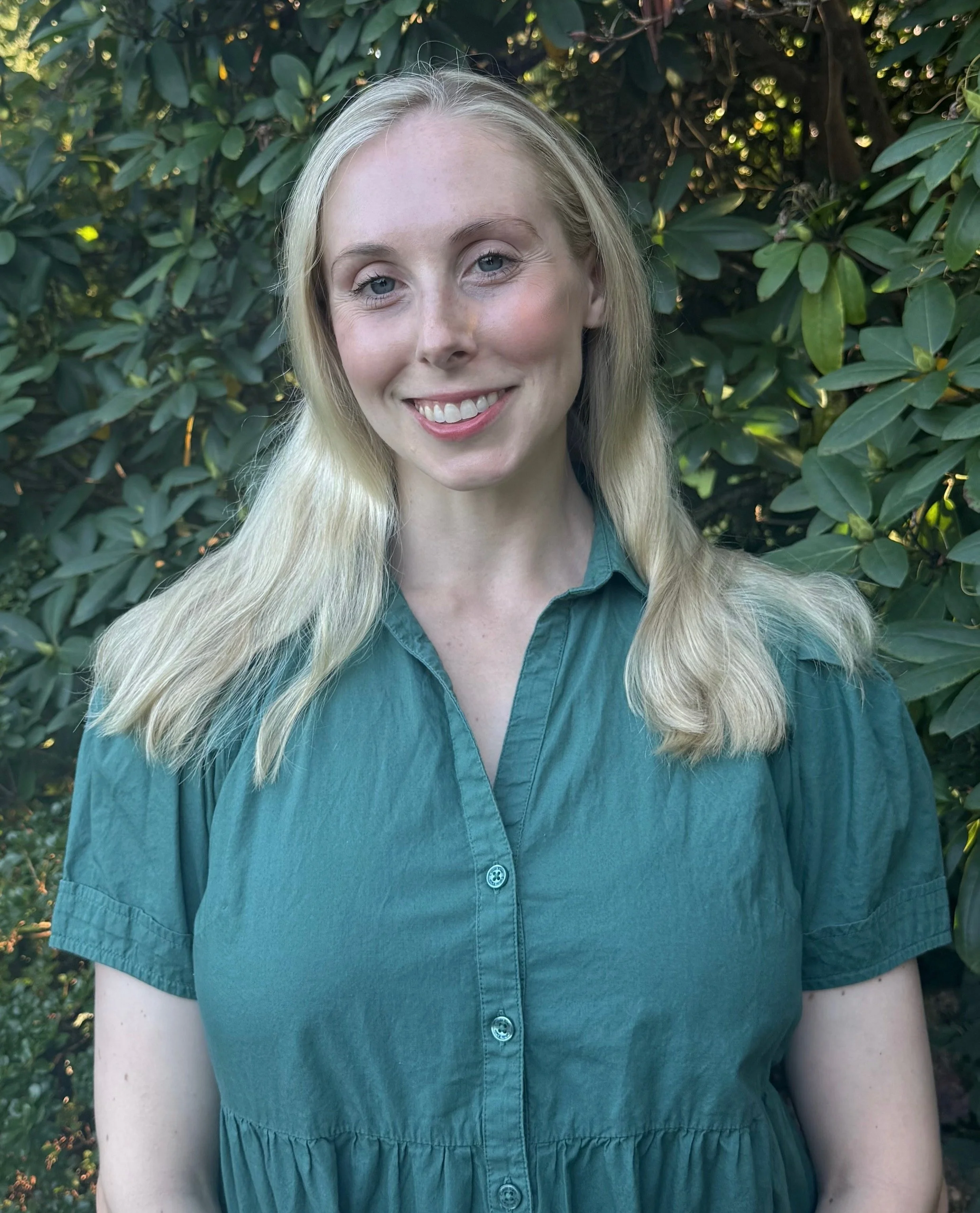 Close-up of a smiling blonde woman wearing a teal button-up shirt, standing outdoors in front of green leafy bushes.