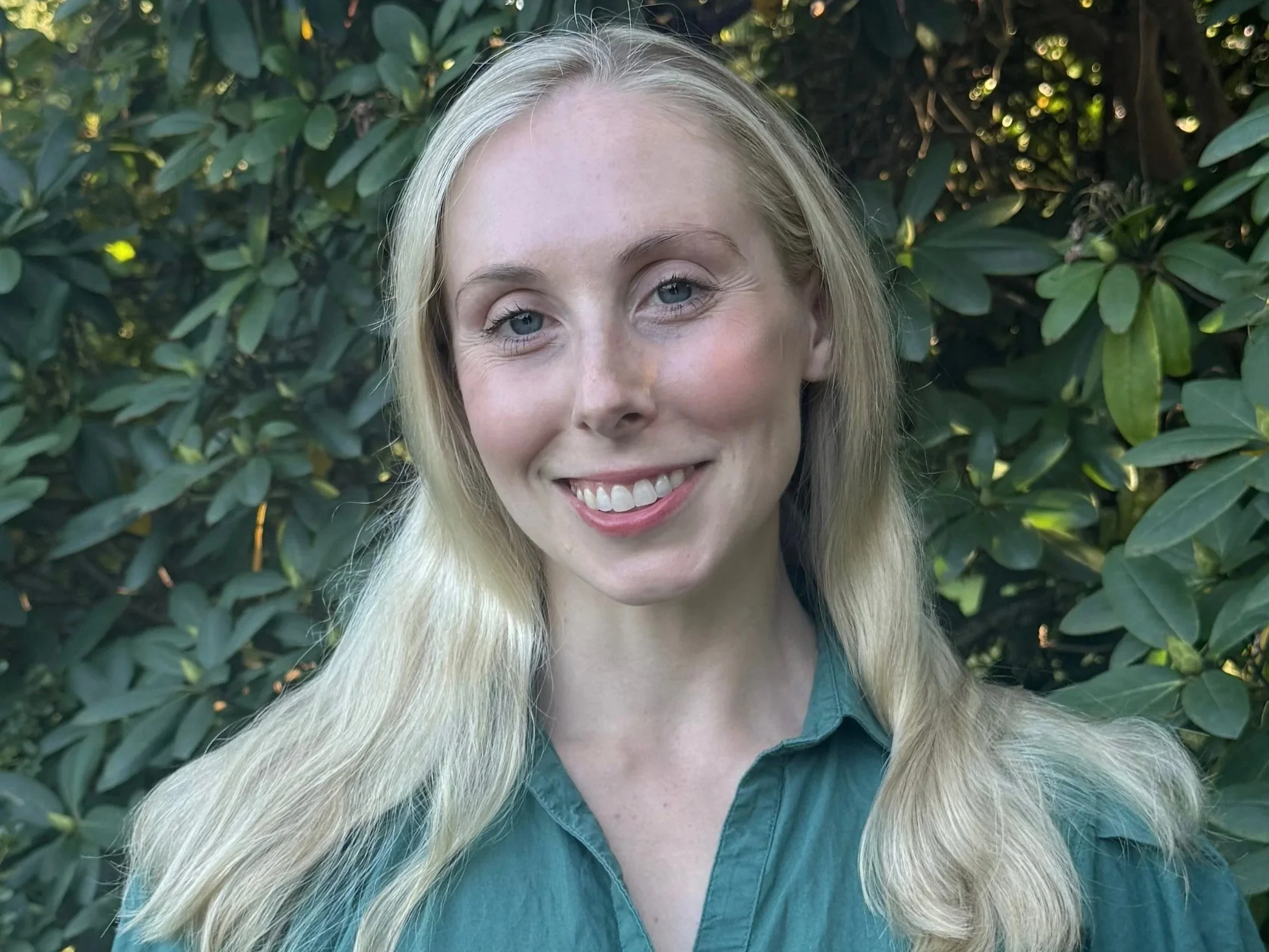 A woman with blonde hair and blue eyes smiling outdoors in front of green leafy bushes.