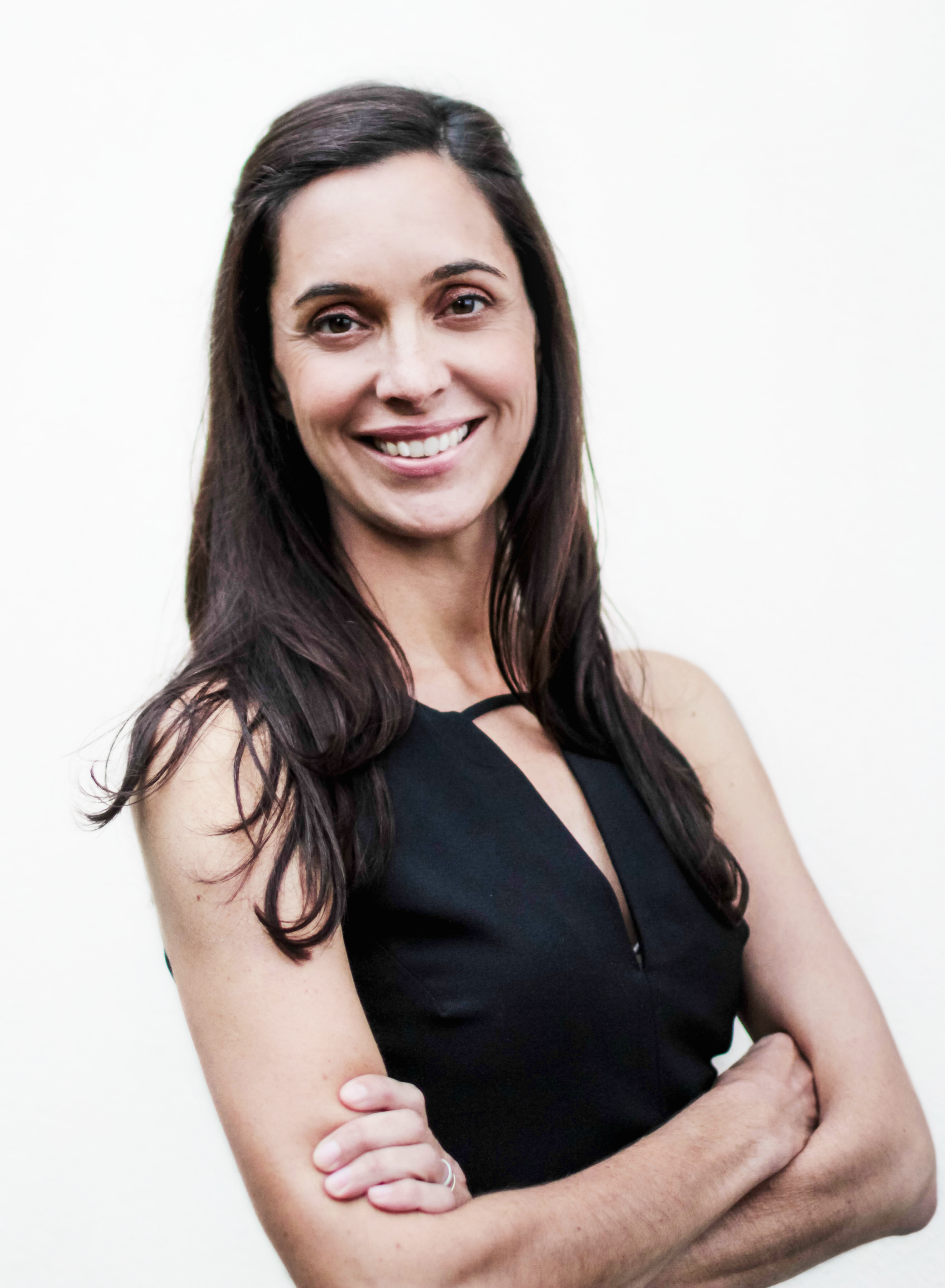 A woman with long dark hair smiling, wearing a black sleeveless dress, standing against a plain white background.