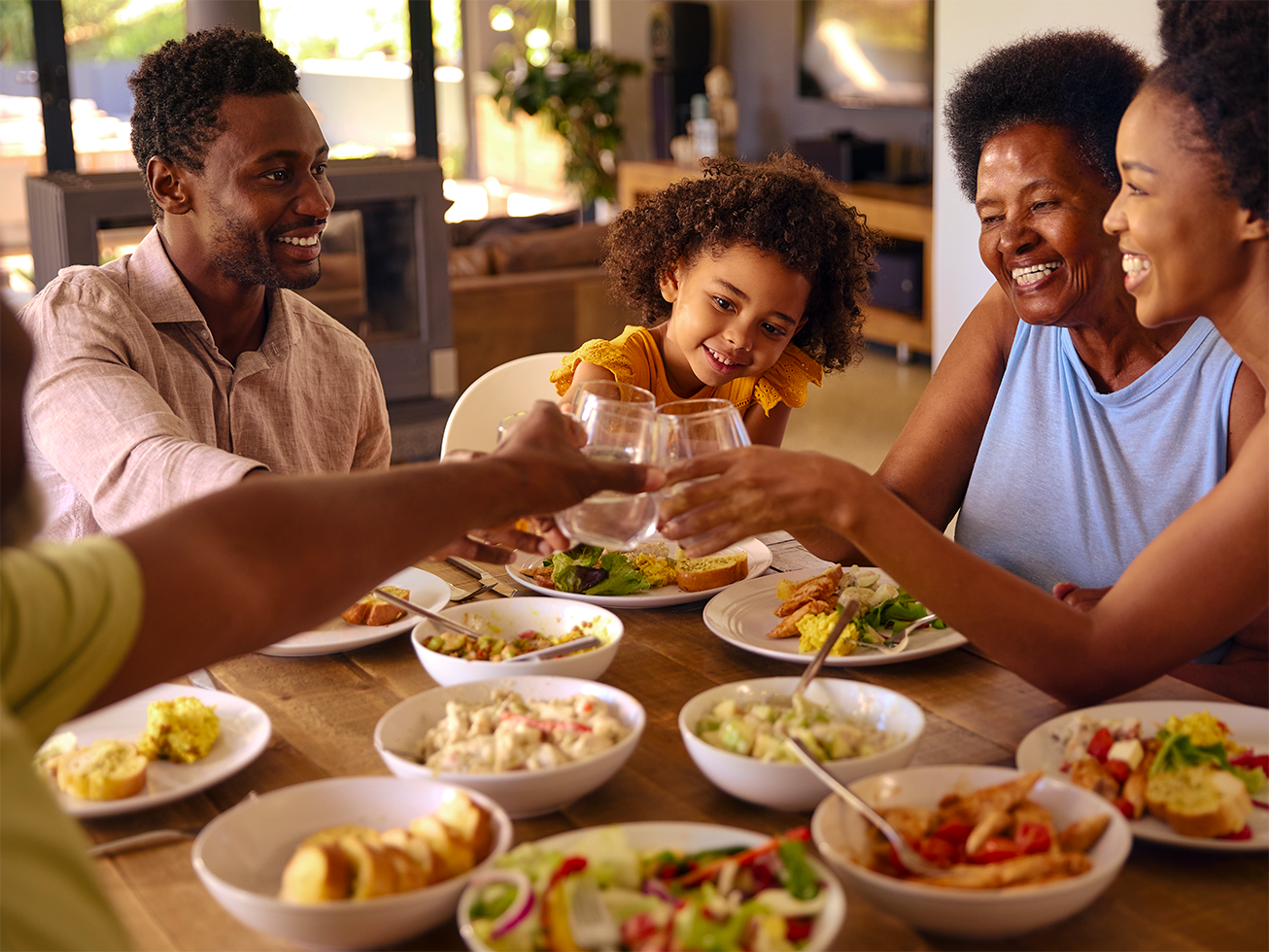Family of four having dinner together, toasting with glasses, with various dishes on the table, in a cozy home setting.