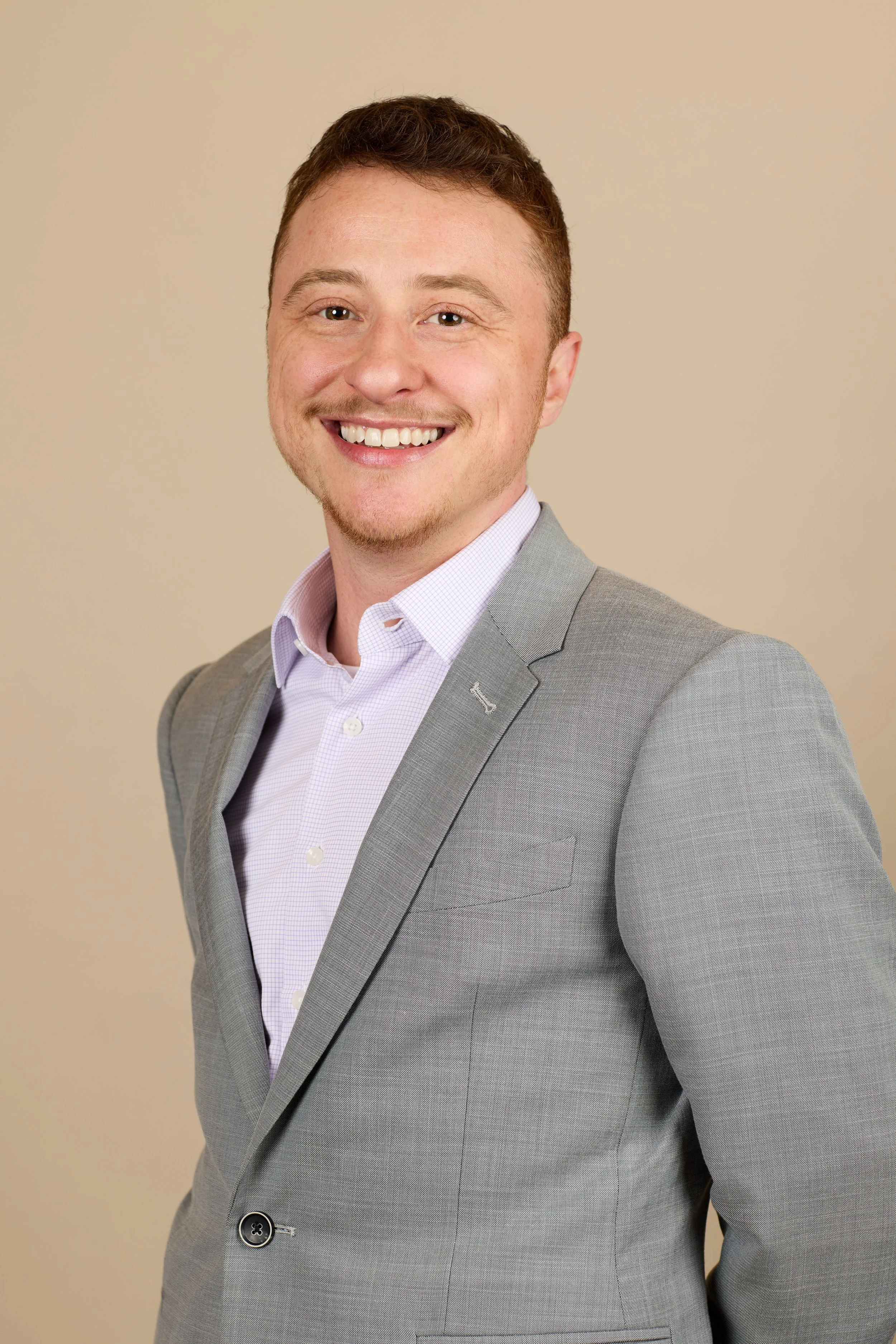 Portrait of a young man with short brown hair, light skin, smiling, wearing a light purple shirt and a gray suit jacket, against a beige background.