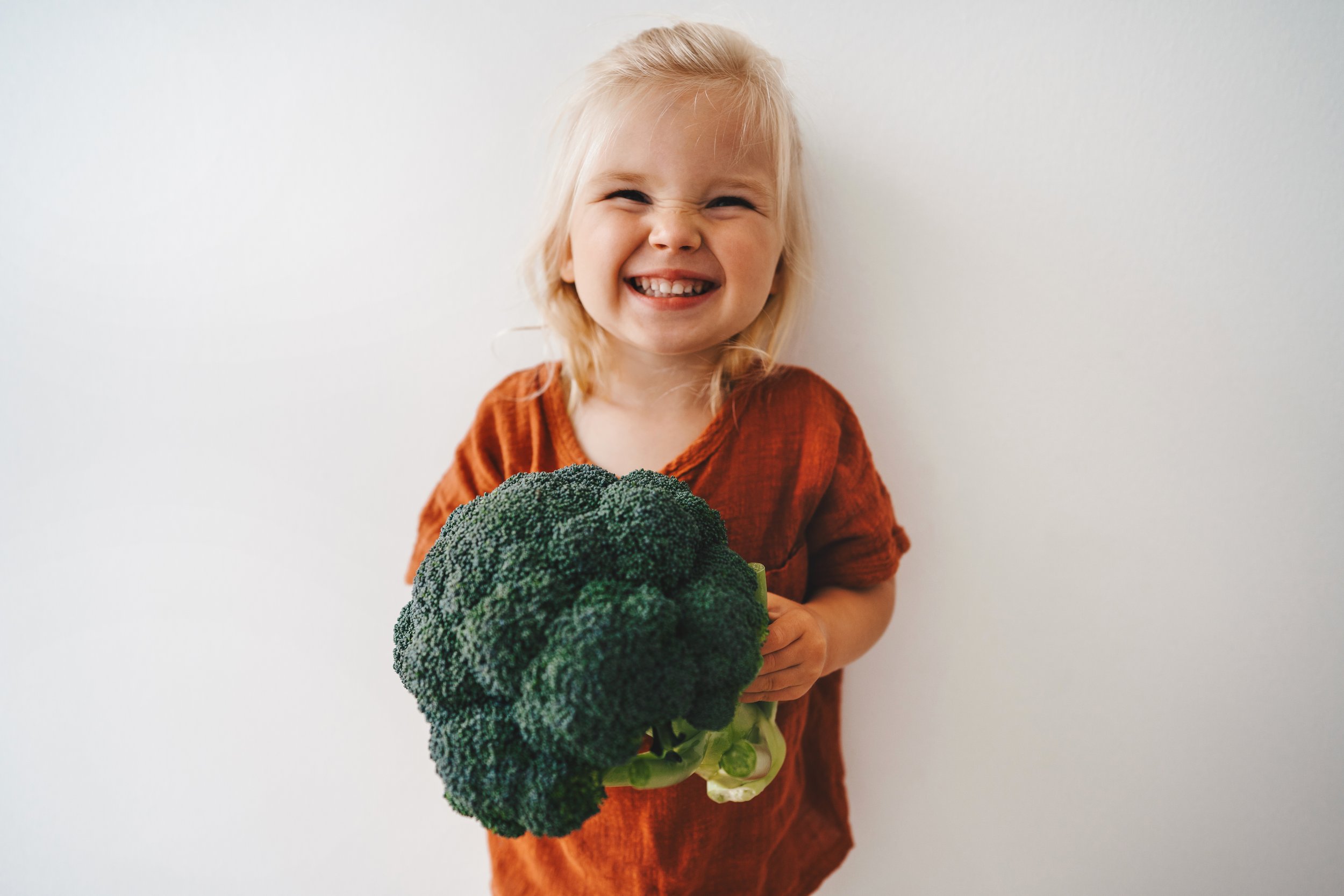 A young girl with blonde hair and a wide smile, holding a large head of broccoli in front of a plain white background.