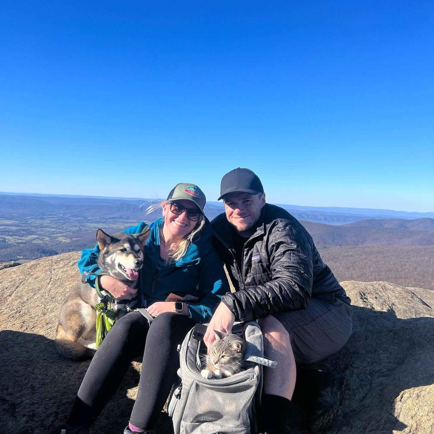 A smiling couple and their dog and cat sitting on a mountain peak with scenic mountains and clear blue sky in the background.