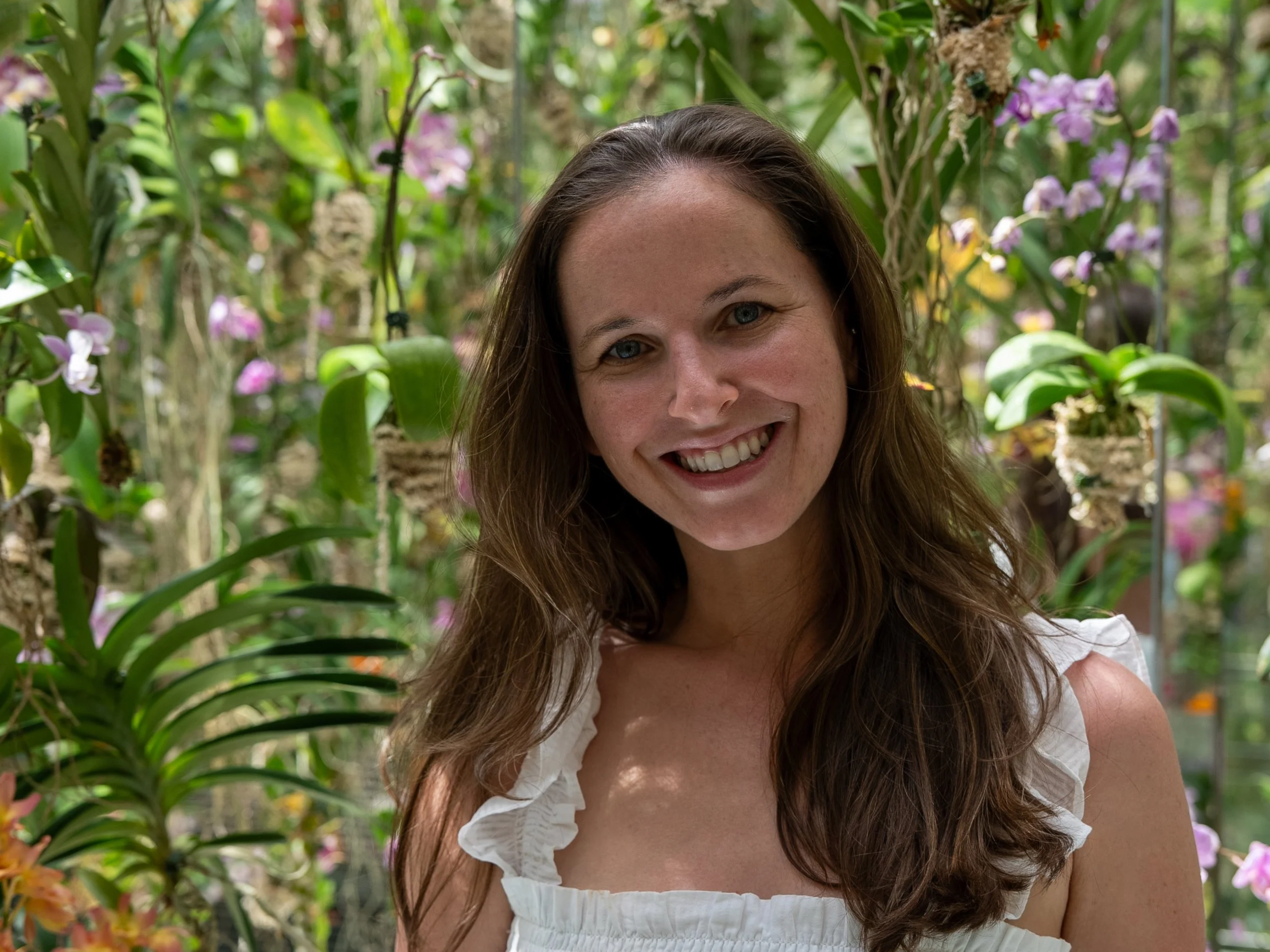 A woman with long brown hair smiling in a lush garden with purple and yellow flowers.