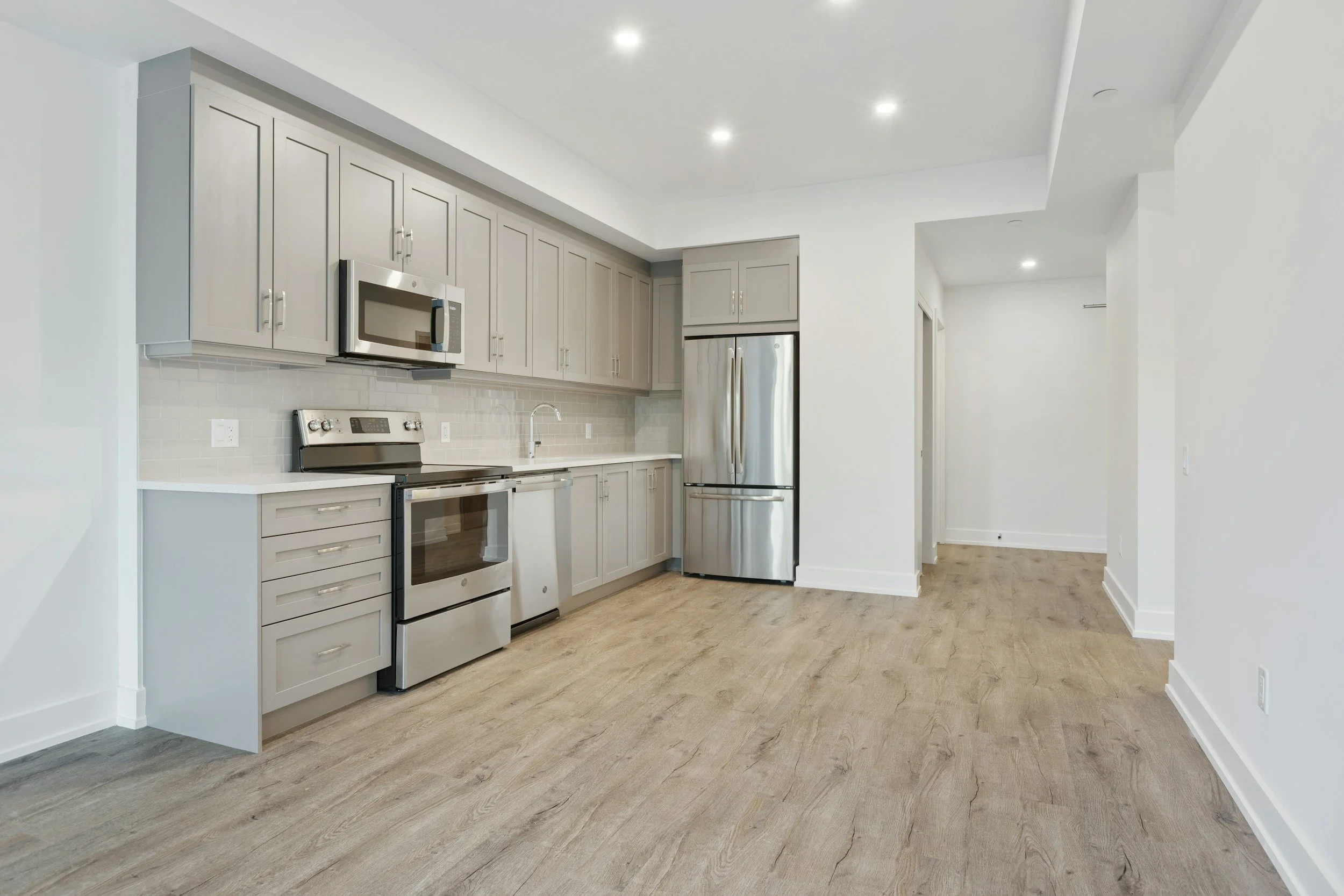 Modern kitchen with gray cabinets, stainless steel appliances, wooden floors, white walls, recessed ceiling lights, and minimal decor.