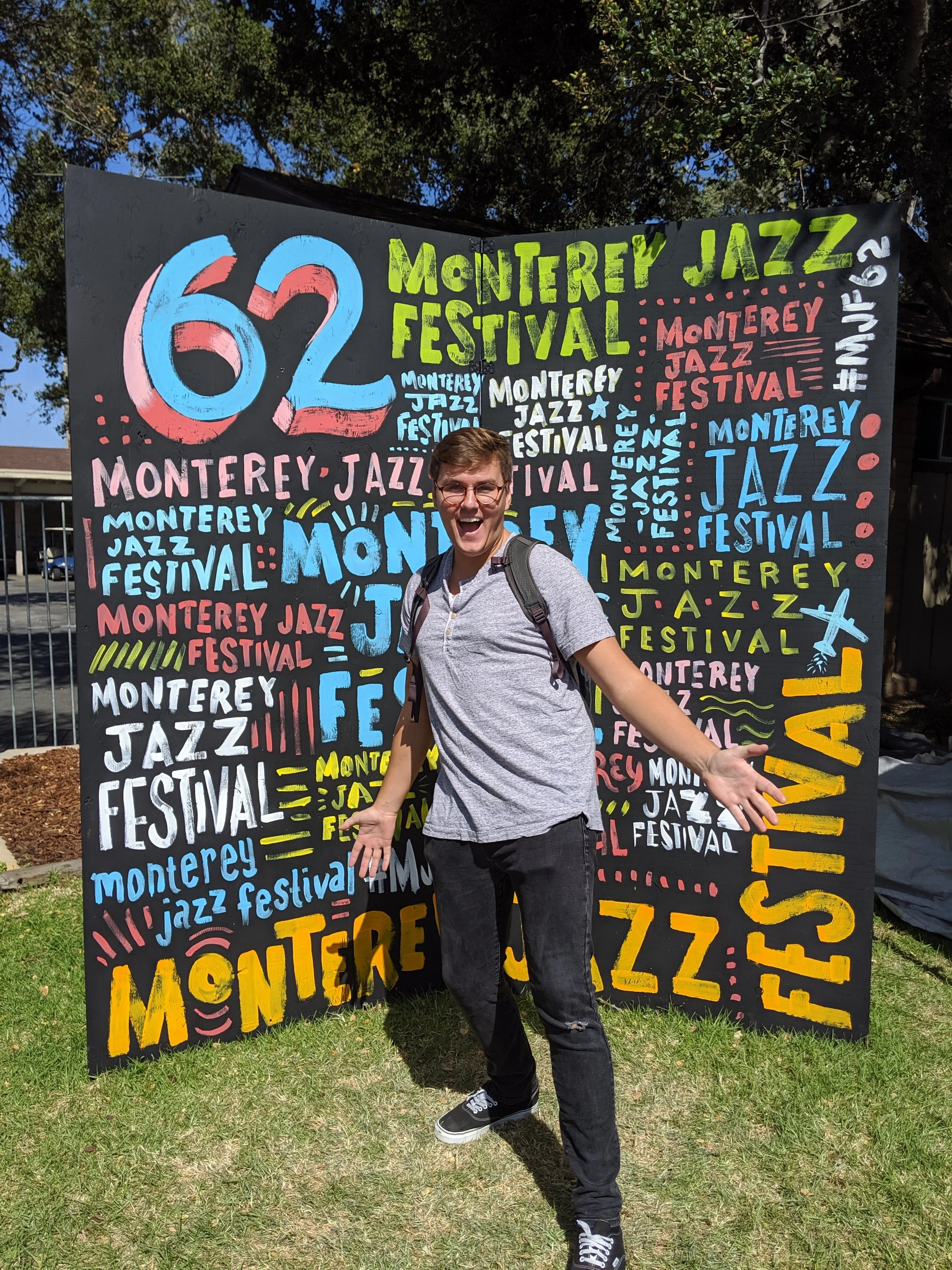 A man with glasses and a backpack standing in front of a colorful banner for the Monterey Jazz Festival, smiling with arms outstretched.