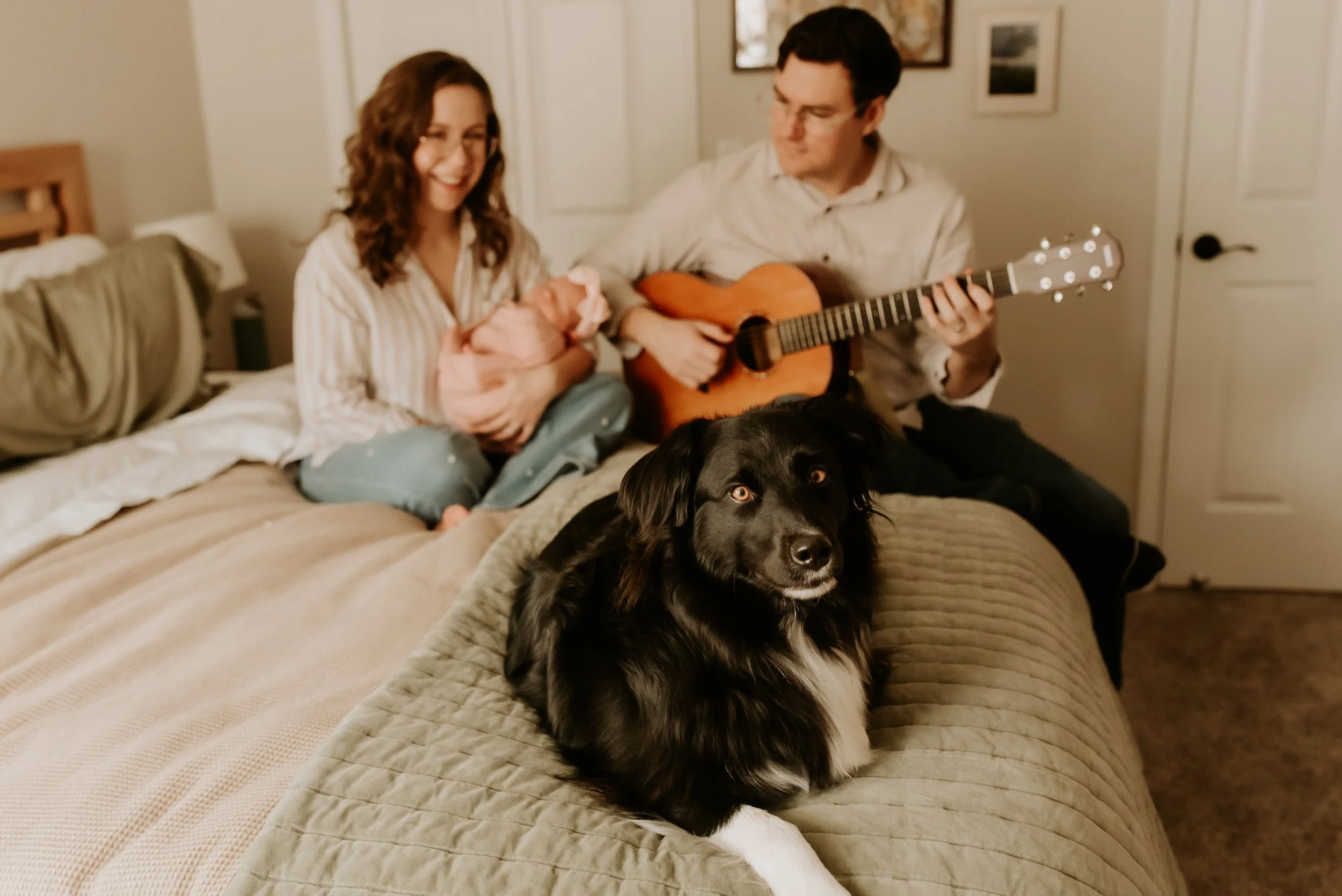 A woman smiling while sitting on a bed next to a man playing acoustic guitar, with a black and white dog lying in the foreground.