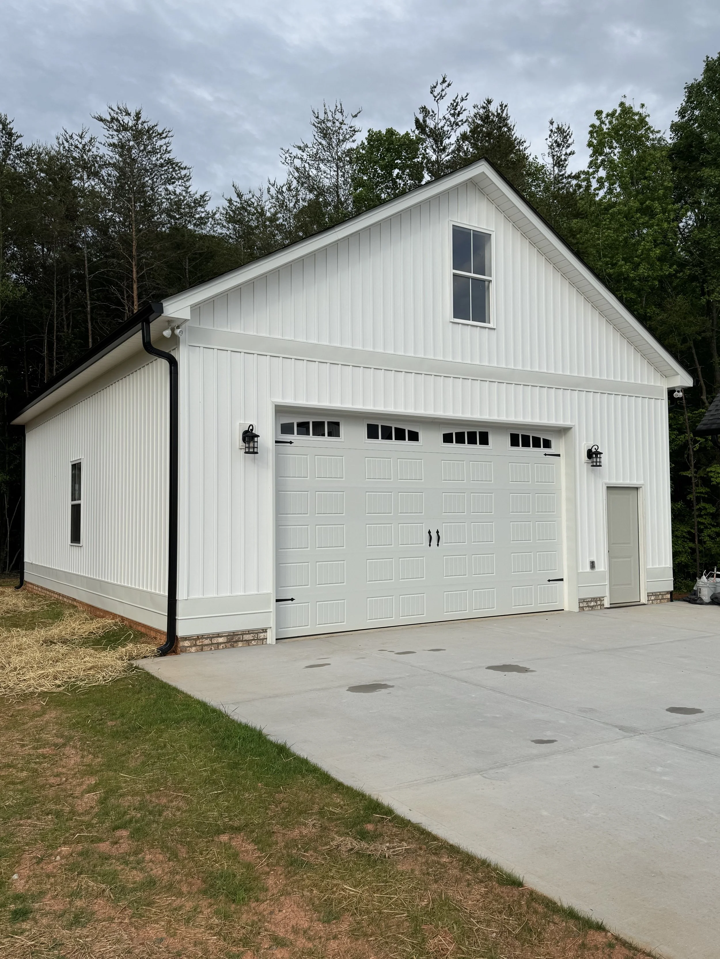 White garage with two doors, a small side door, black outdoor wall lights, and a window on the upper level, surrounded by trees and a cloudy sky.