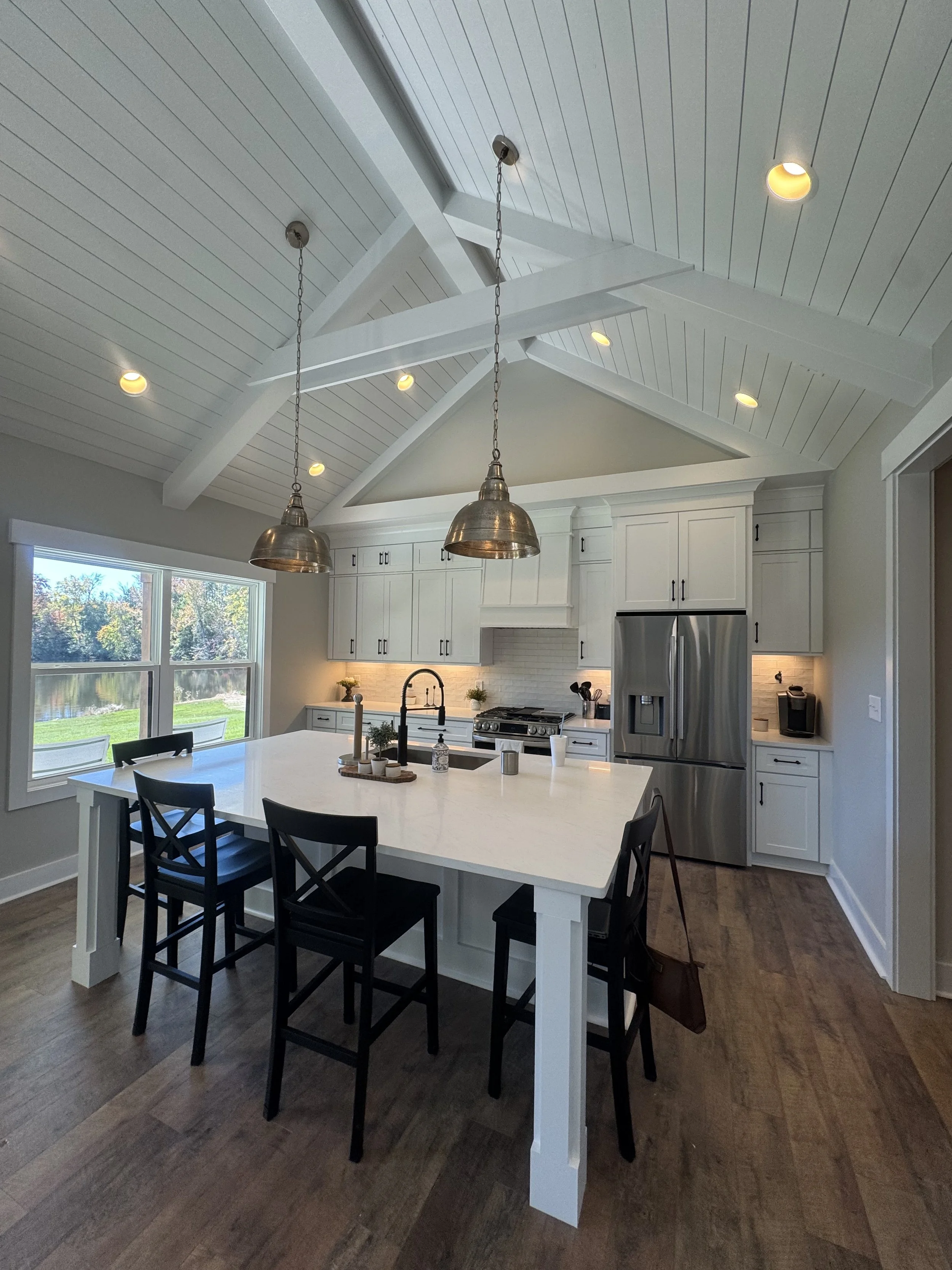 A modern kitchen with a white island countertop and black chairs, stainless steel appliances, white cabinets, large window showing outdoor trees, pendant lights hanging from a white beamed ceiling, and hardwood flooring.