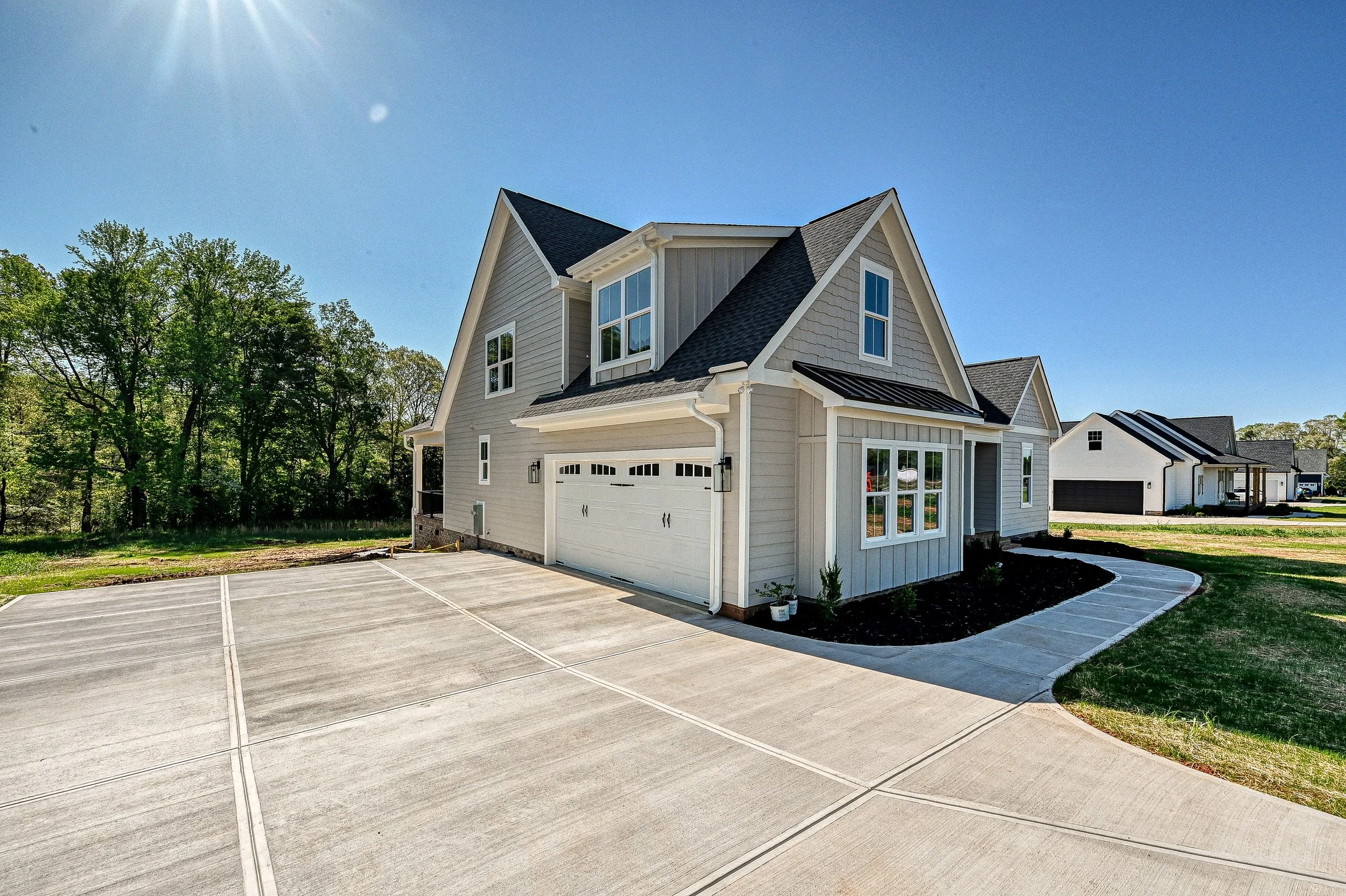 A newly built two-story house with a garage, concrete driveway, and well-maintained yard, surrounded by trees and other houses.