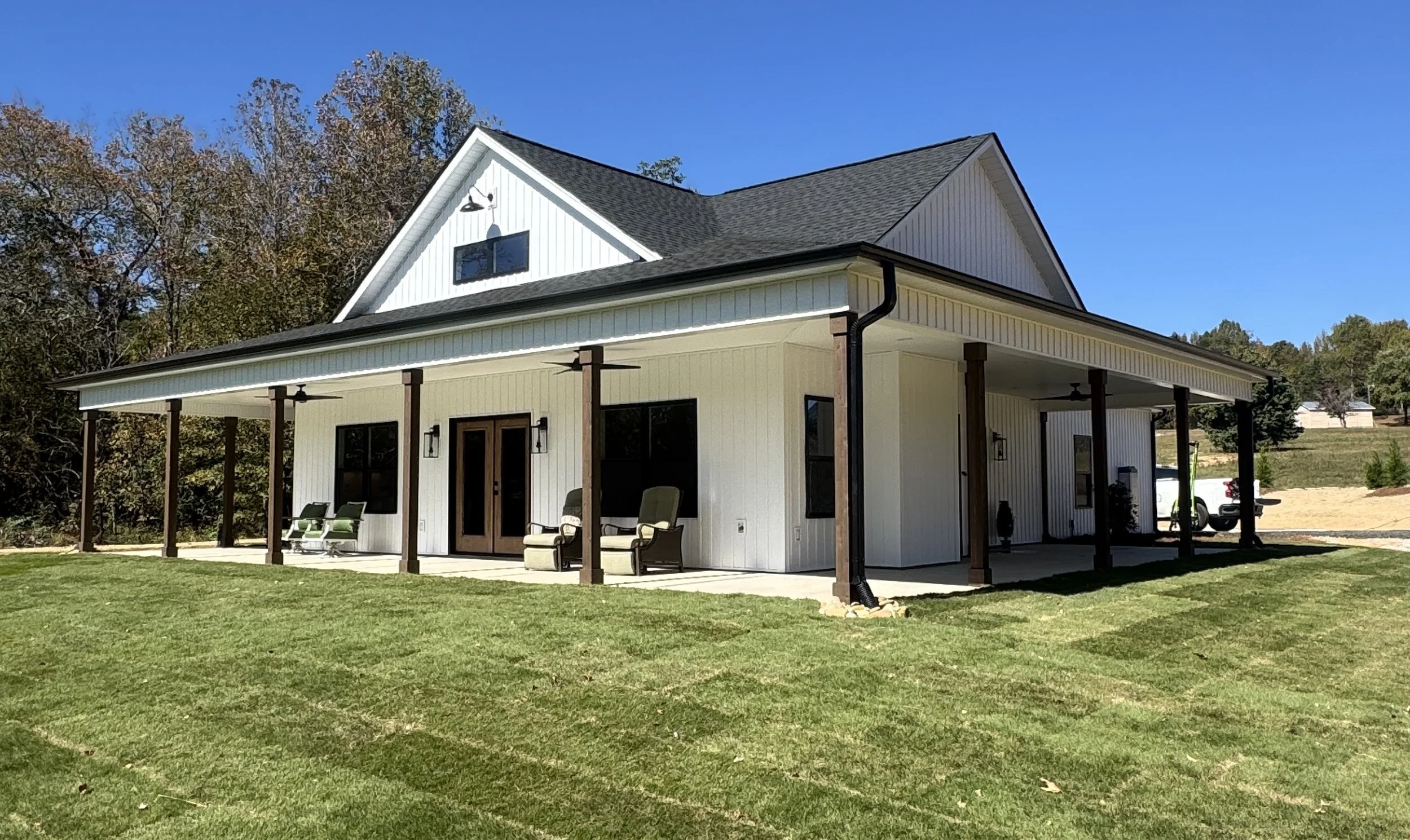 A modern two-story house with white siding and a dark gray roof. The house features a large covered porch with outdoor seating, ceiling fans, and a grassy yard. There are trees in the background and a clear blue sky.