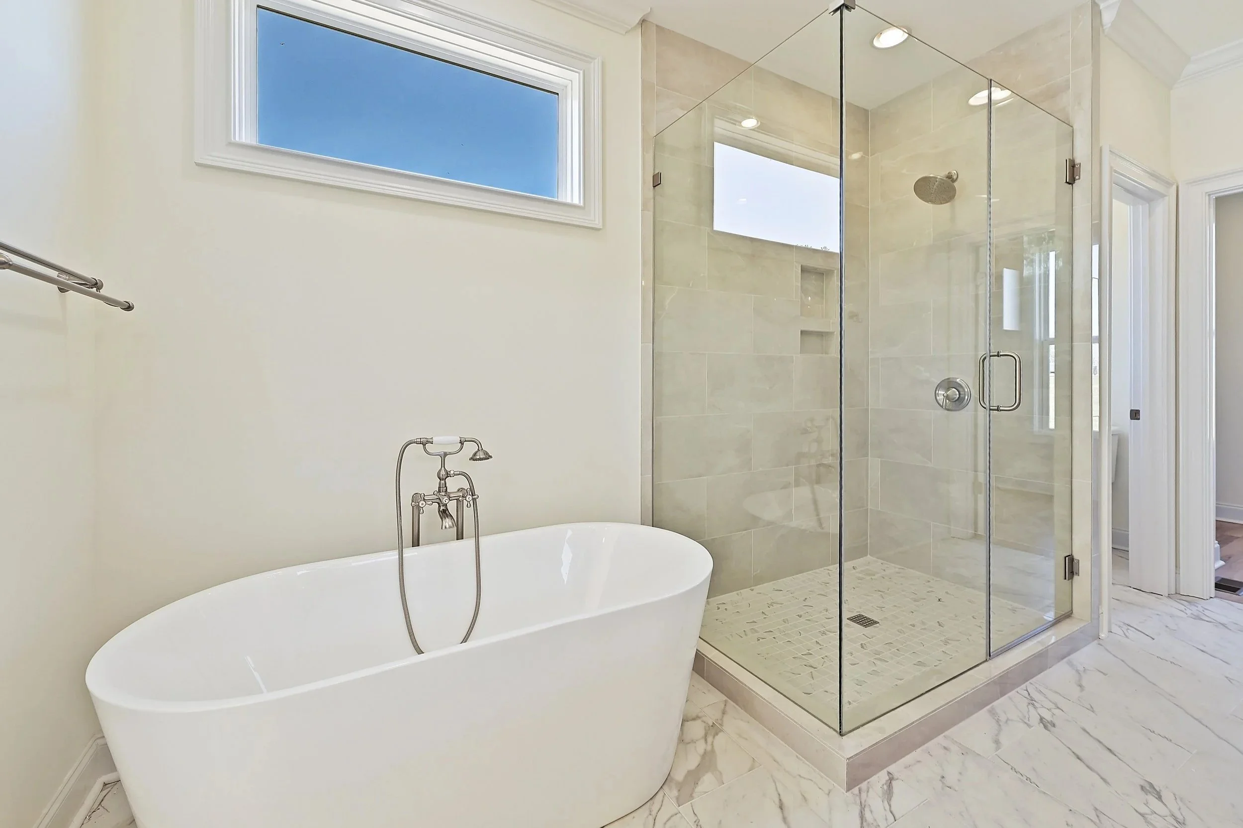A modern bathroom featuring a white freestanding bathtub with a vintage style faucet, a glass-enclosed shower with beige tiles, a small niche in the shower, a marble floor, a large window, and marble flooring.