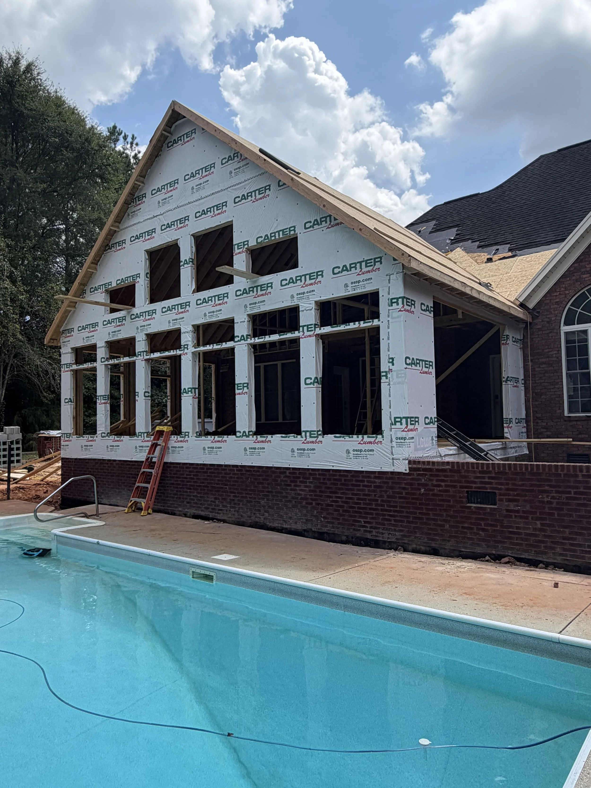 A house under construction with a partially built upper floor, surrounded by a brick wall, with a swimming pool in the foreground under a partly cloudy sky.