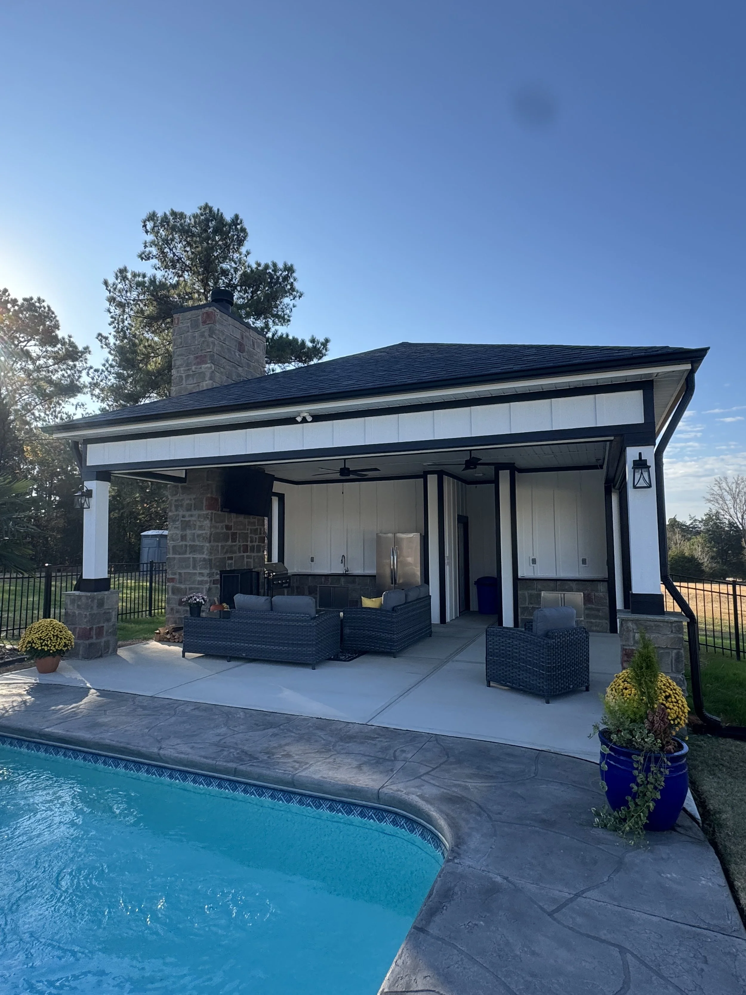 A backyard with a swimming pool, patio furniture, potted plants, a covered patio area, a brick chimney, and tall trees in the background under a clear blue sky.