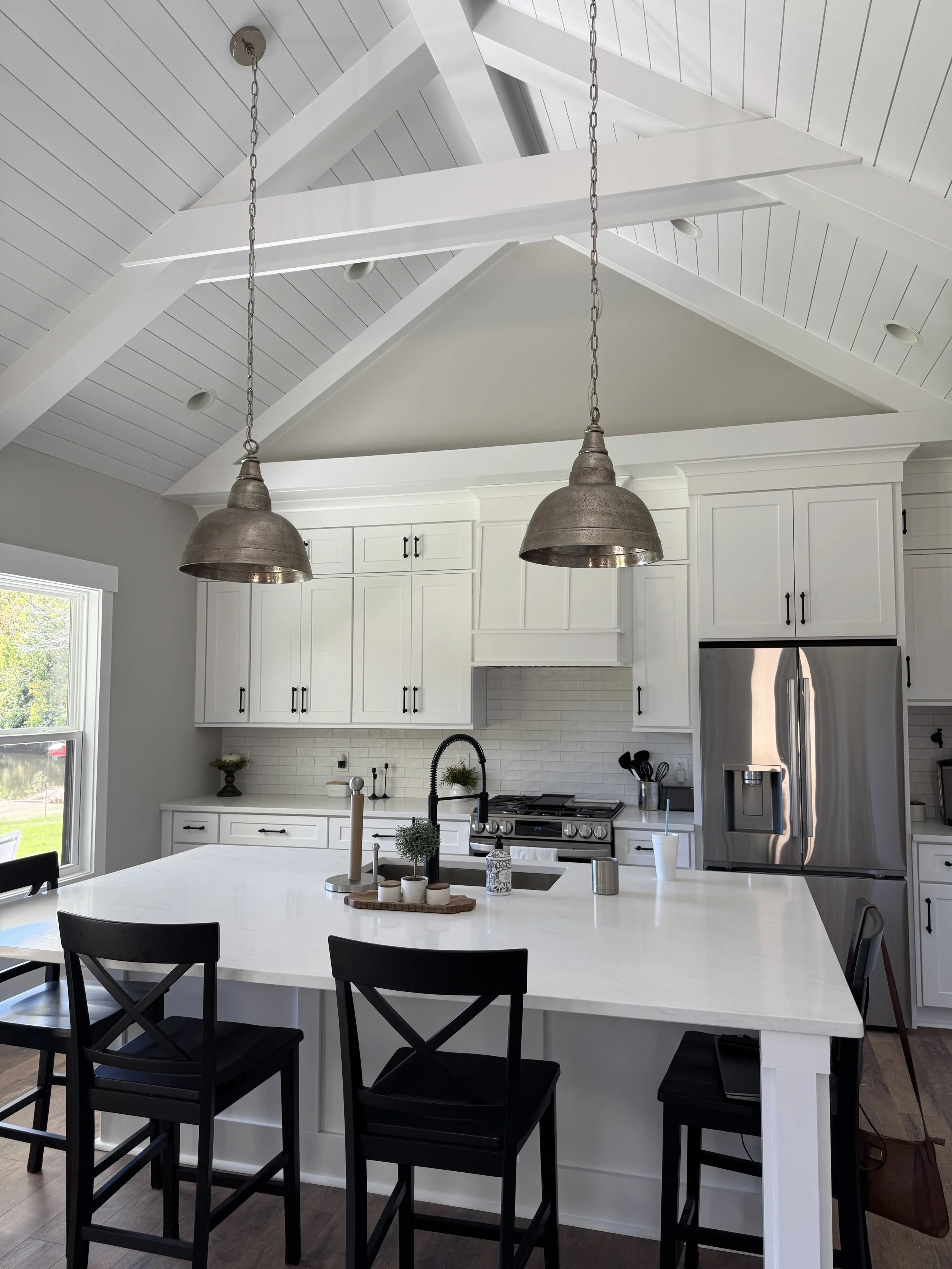 A modern kitchen with white cabinetry, stainless steel appliances, a large island with a white countertop, and black chairs around the island. Two industrial pendant lights hang over the island. There is a window on the left side and a door with a window on the bottom left corner.