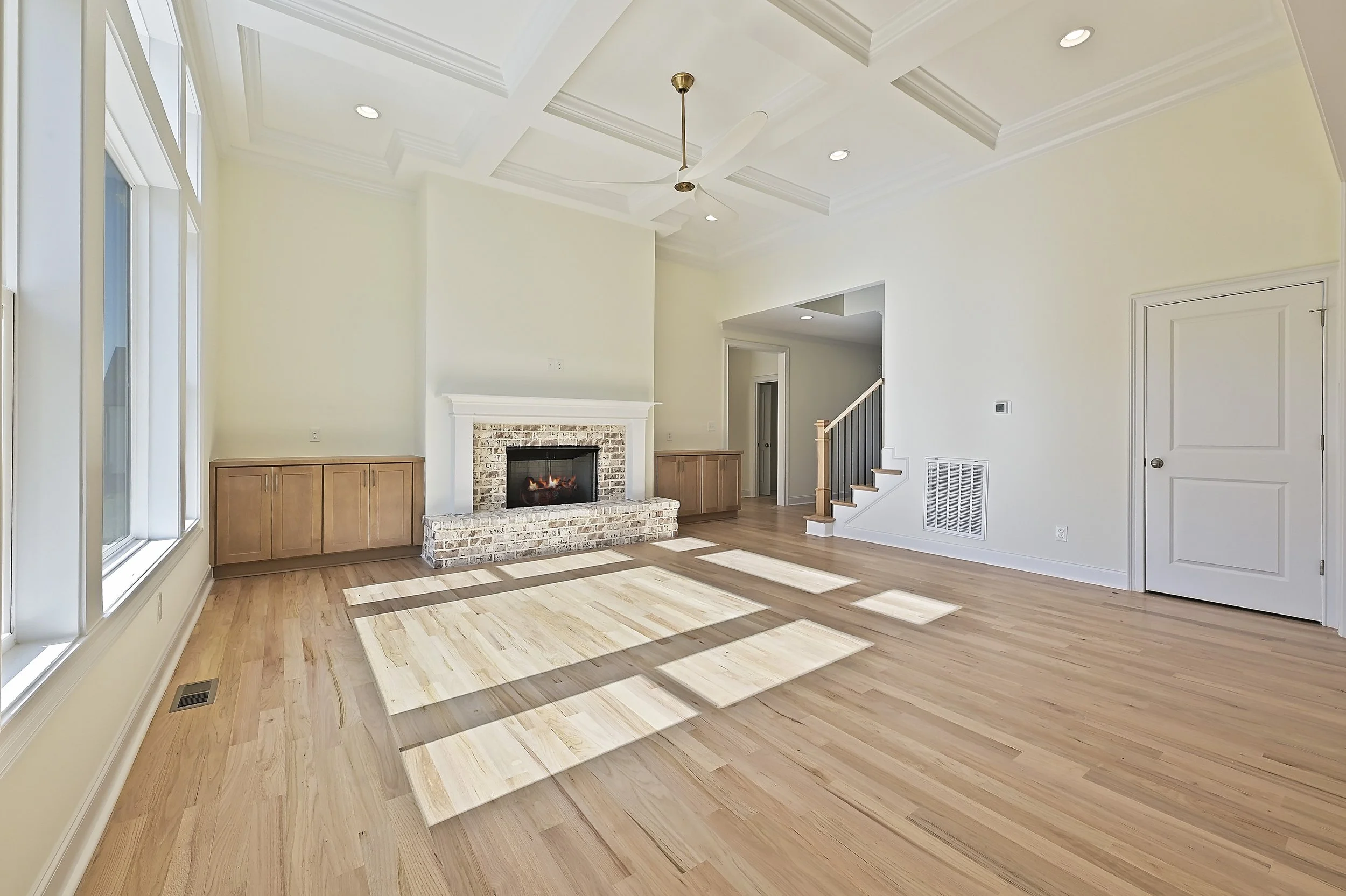 Empty living room with light wood flooring, white walls, a brick fireplace with wooden cabinets, high ceiling with a fan, large windows on the left, and a staircase with a wooden handrail on the right.