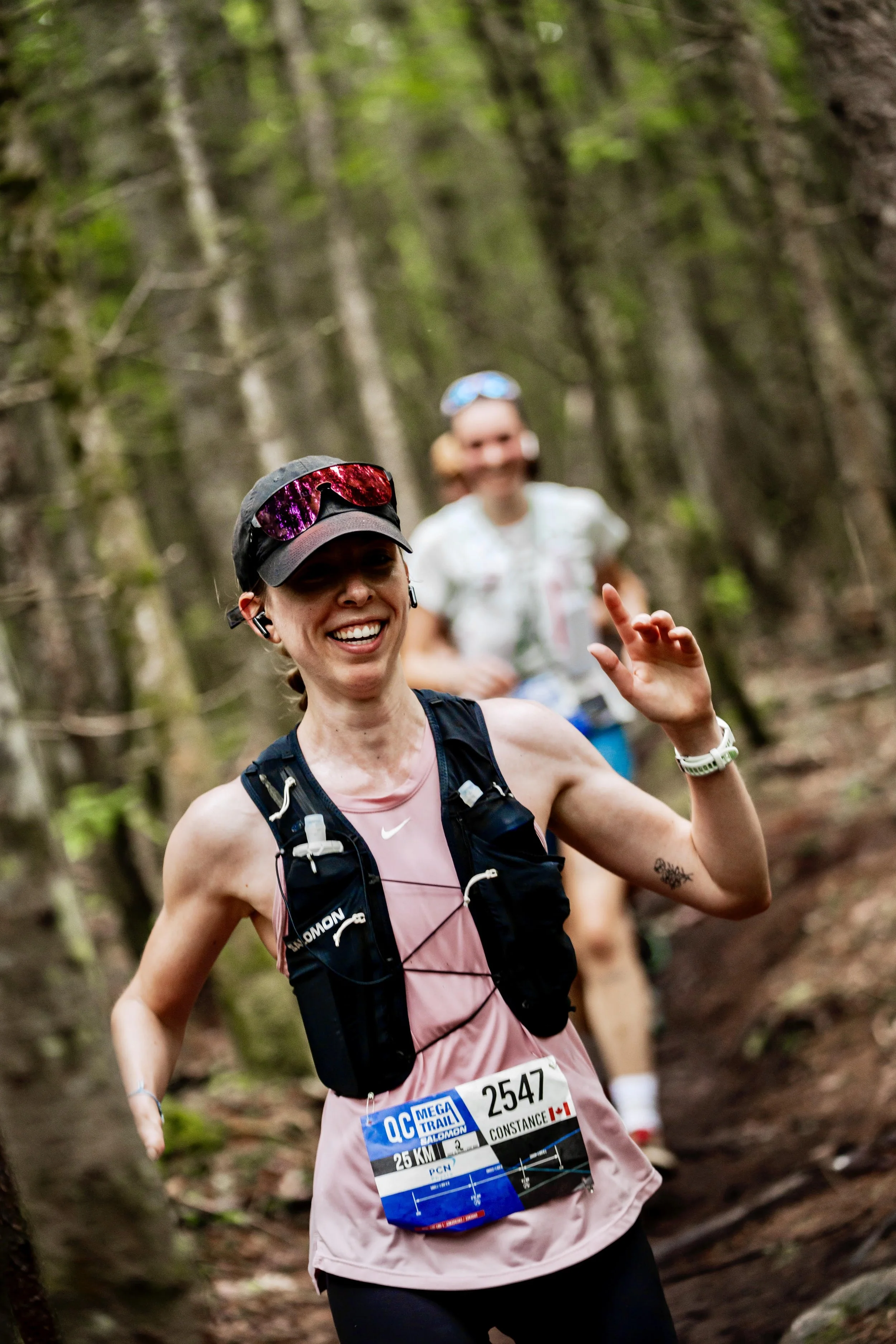 Smiling woman running in a forest trail during a race, wearing a pink tank top, running vest, race bib, sunglasses, and Bluetooth earbuds, with another runner blurred in the background.