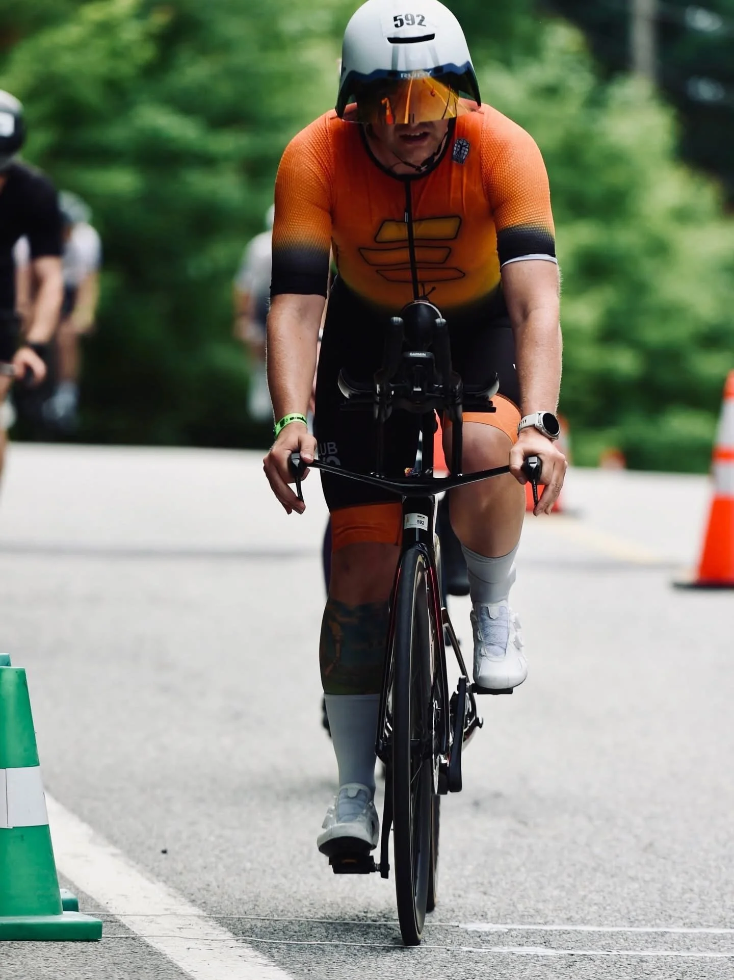 A cyclist in orange cycling gear and a helmet riding a bike through a marked lane, with cones and trees in the background.