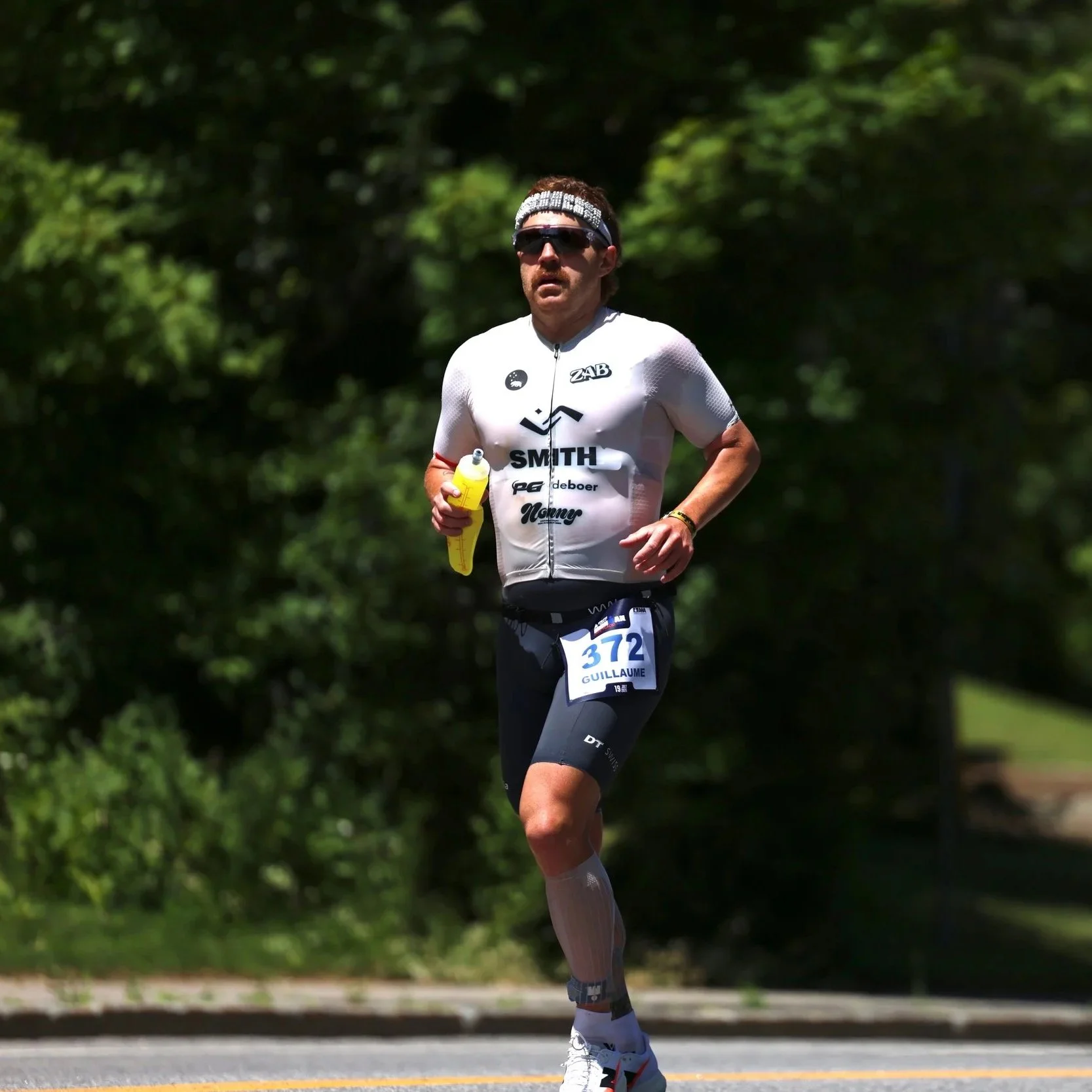 Male runner in white athletic gear, sunglasses, and headband running on a tree-lined outdoor path, holding a yellow water bottle.