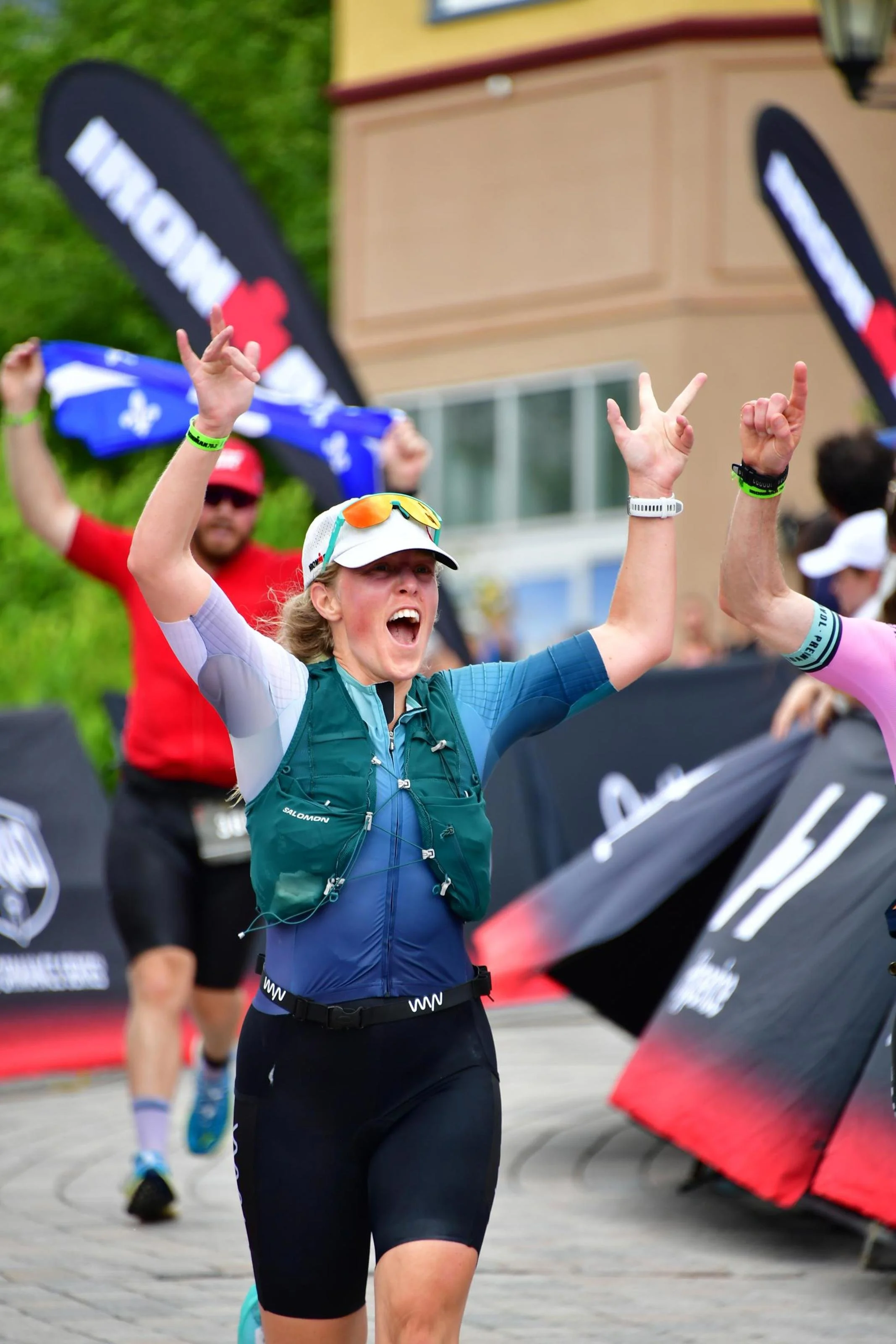 A woman participating in a running race, raising her hands in celebration near a finish line with other runners and spectators in the background.