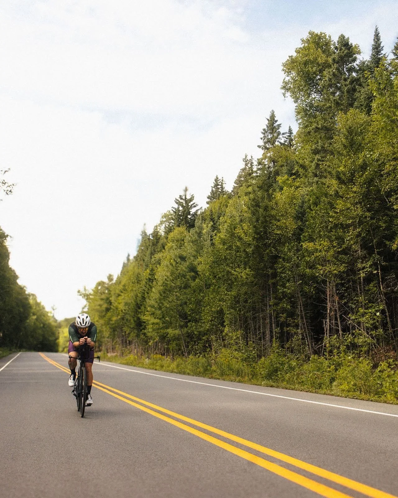 A person riding a bicycle on a rural road surrounded by green trees and foliage under a partly cloudy sky.