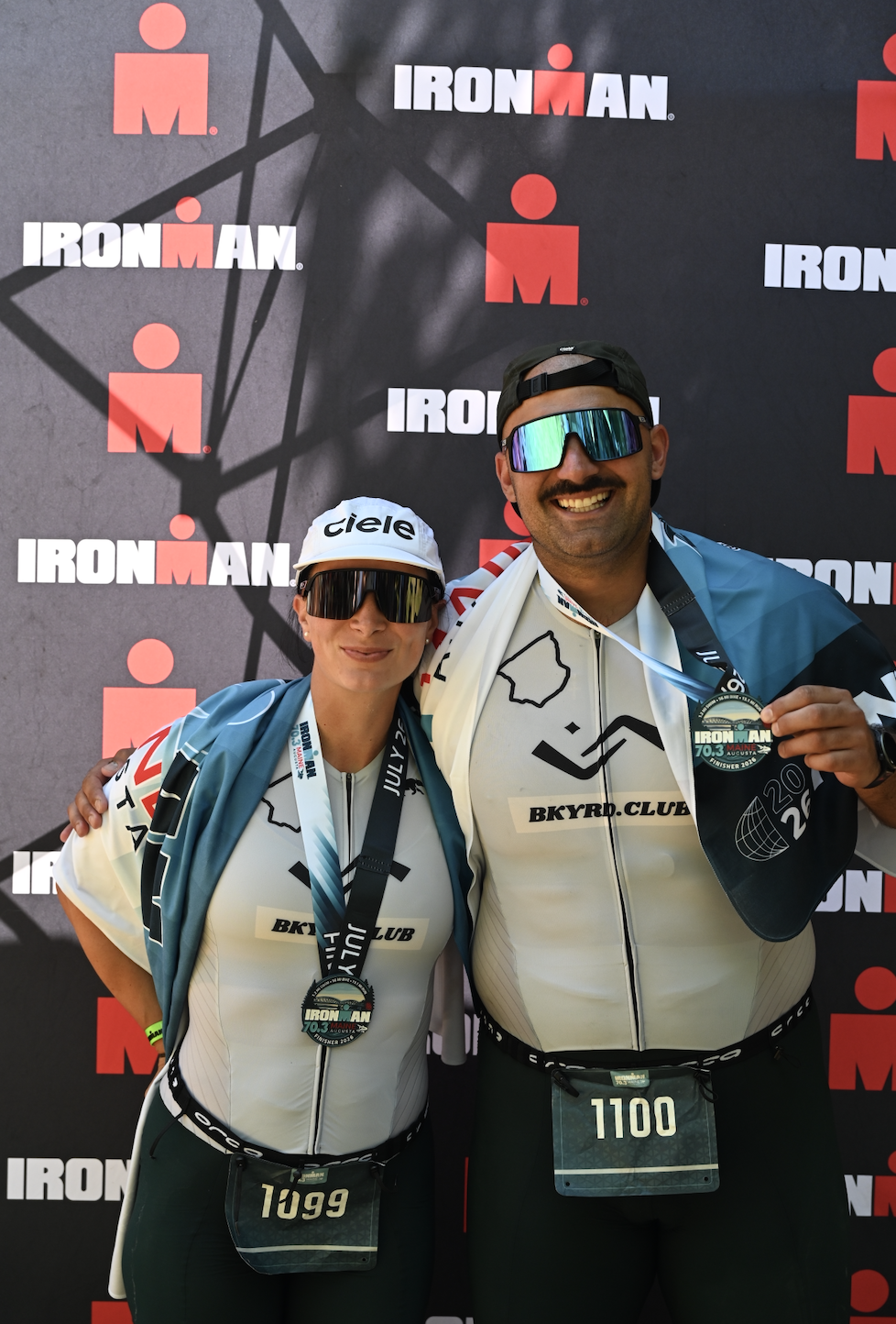 Two athletes wearing medals and athletic gear standing in front of an Ironman backdrop, celebrating their participation in the event.