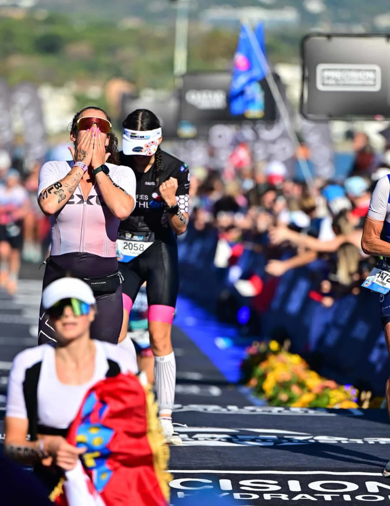 Two women running in a marathon, one with hands covering face and the other celebrating with arm raised, with a crowd and colorful flowers along the route.