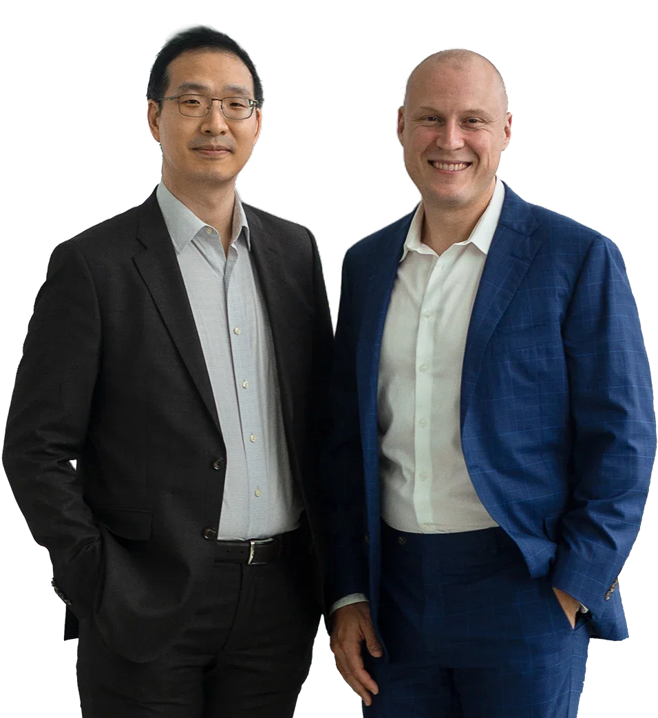 Two men in suits standing side by side, smiling, with one wearing glasses and a black suit and the other bald and wearing a blue suit.