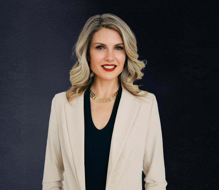 Professional woman with blonde, wavy hair wearing a white blazer and black top, smiling against a dark background.