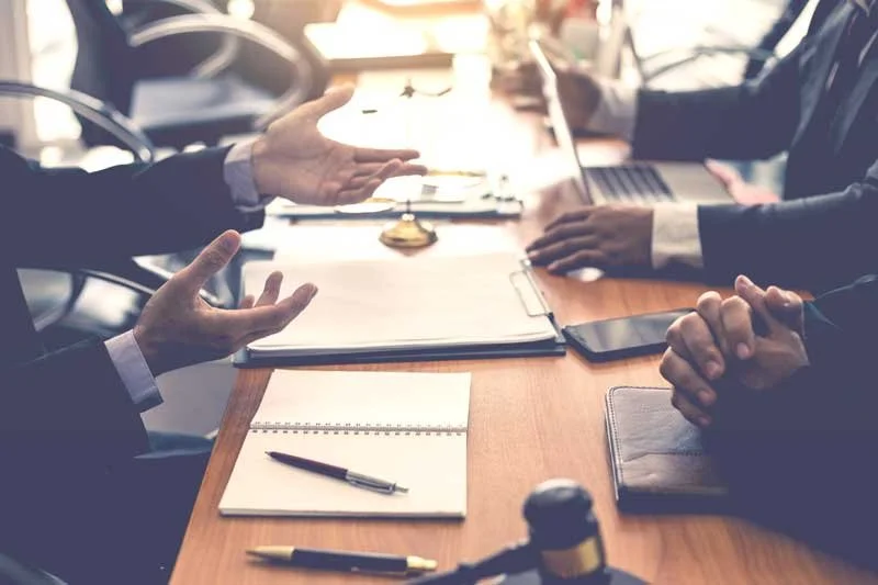 Business meeting with multiple people sitting at a wooden conference table, engaging in discussion. Items on the table include documents, notebooks, pens, a smartphone, and a laptop.