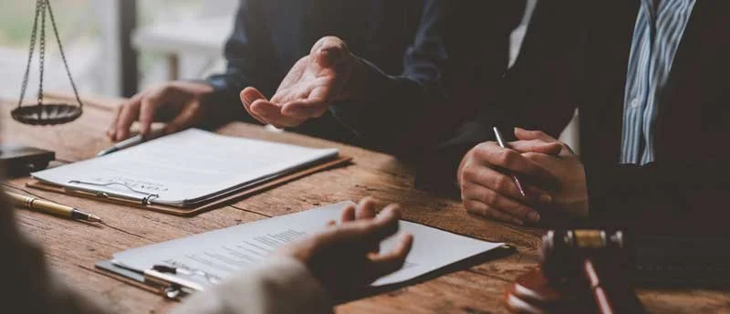 Two people in suits having a discussion at a wooden desk with documents, pens, and a gavel.