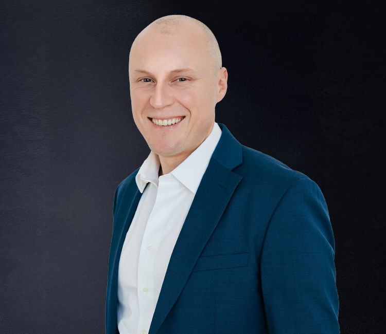 Headshot of a smiling man in a white shirt and blue blazer against a dark background.