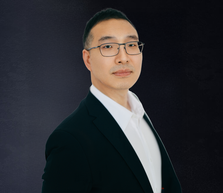 A man wearing glasses, a white shirt, and a black blazer standing against a dark background.