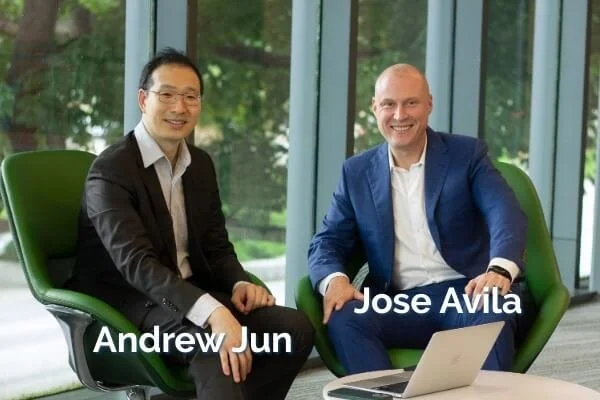 Two men sitting in green chairs in front of large glass windows, smiling at the camera. One is wearing a black suit, the other a blue suit. Labels identify them as Andrew Jun and Jose Avila.