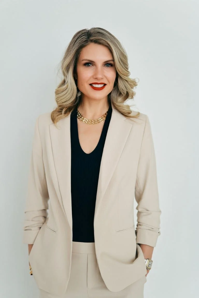 Woman with blonde wavy hair wearing a beige blazer, black top, gold necklace, and watch, standing against a light gray background.