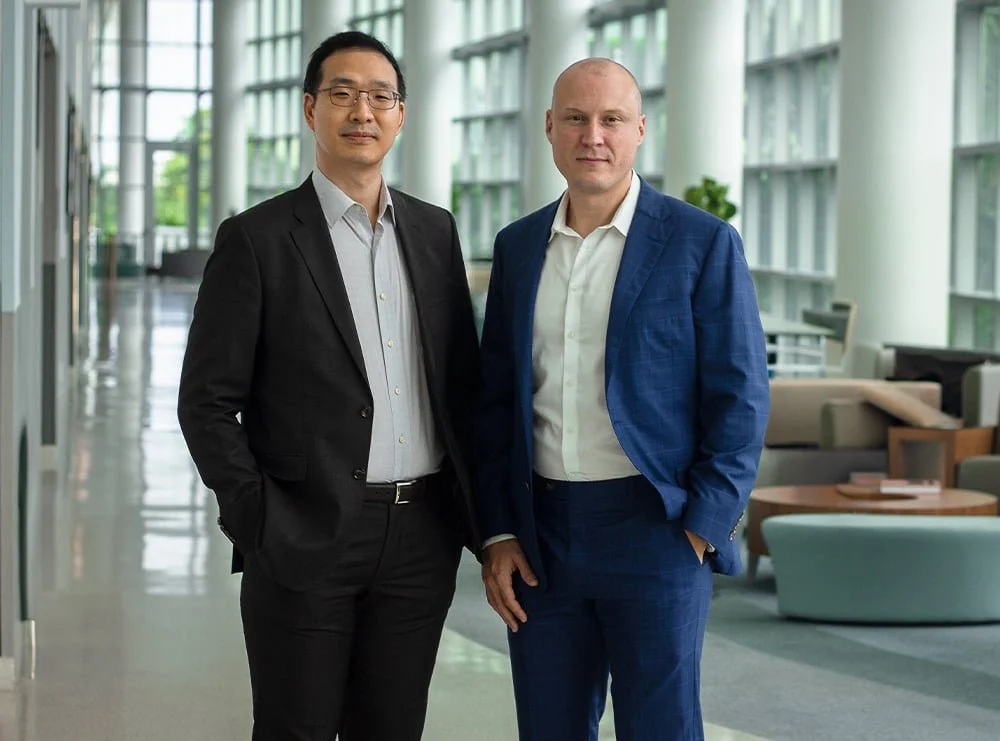 Two men in business suits standing side by side in a modern office lobby with large windows, plants, and seating areas.