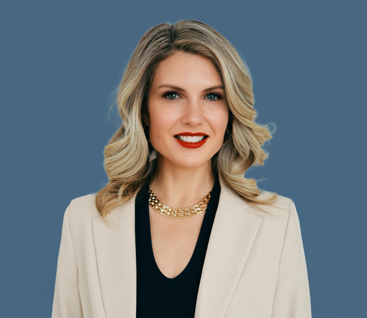 A woman with blonde wavy hair, wearing a black top, a white blazer, a gold chain necklace, and red lipstick, smiling against a blue background.