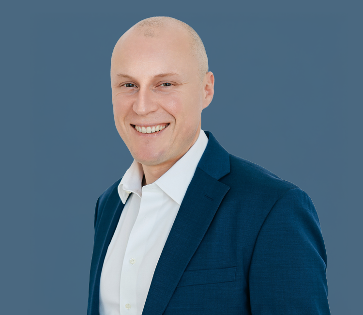 A smiling man in a navy blue suit and white shirt against a solid blue background.