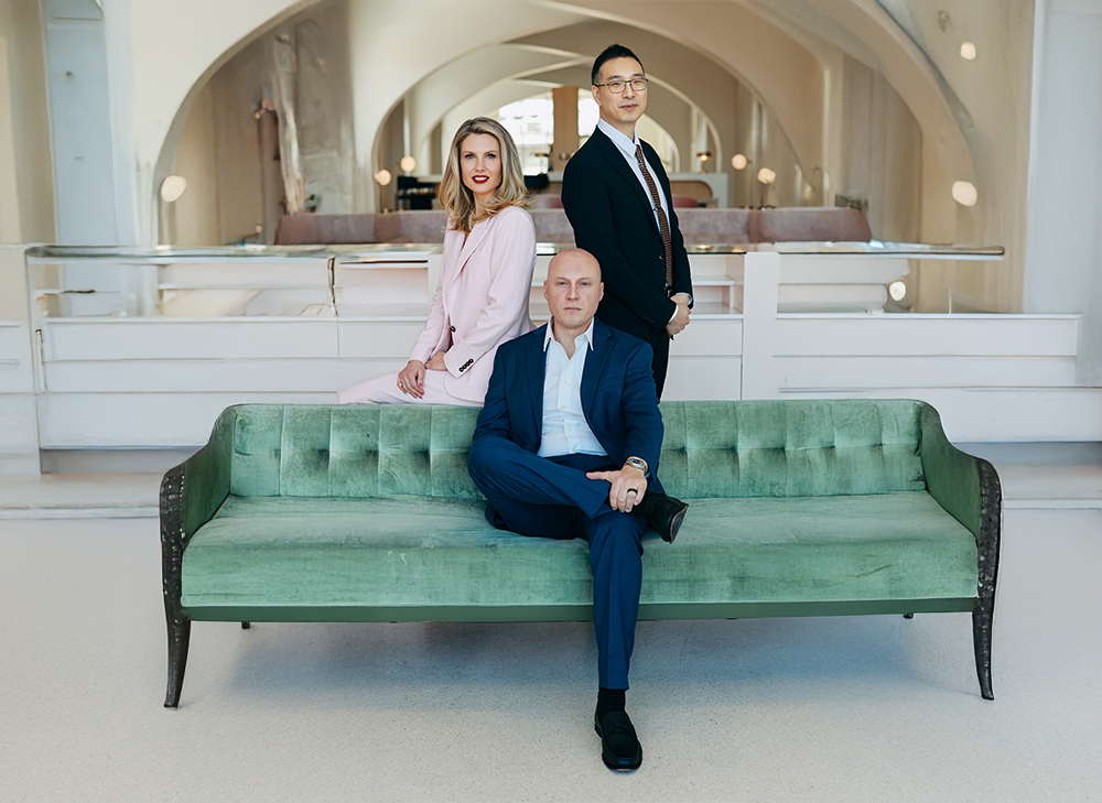 Three professionally dressed people in a modern, elegant interior. One man is sitting on a green velvet sofa with his legs crossed, and two others, a woman in a pink suit and a man in a dark suit, are standing behind him in a well-lit space with curved arches.
