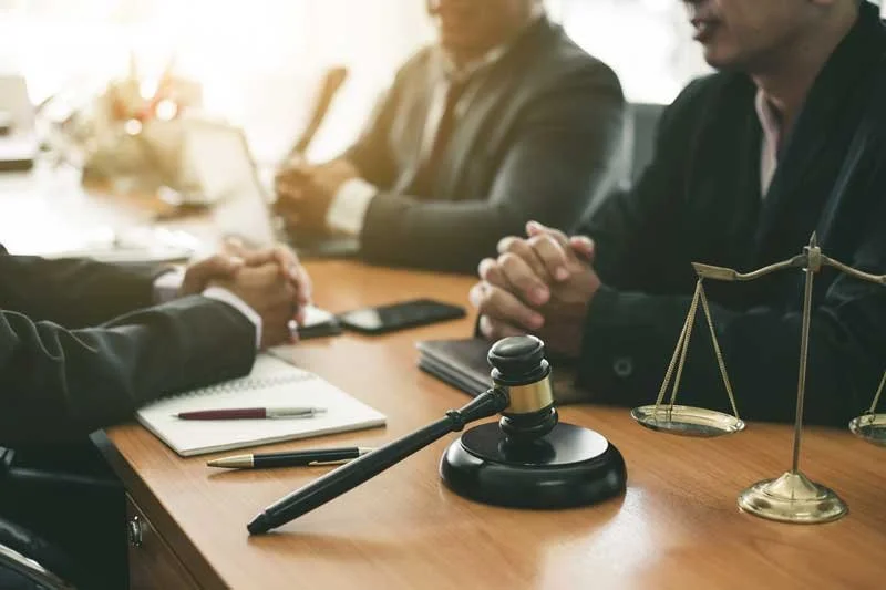 A meeting with three people in business suits, two men with hands clasped, sitting at a wooden table with legal symbols, a gavel, and a scale, during daytime.