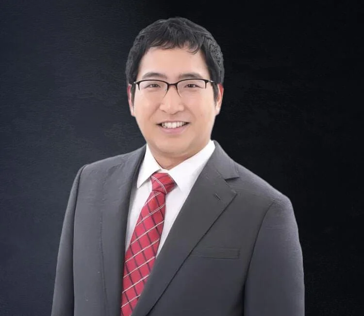 A smiling man in a gray suit, white shirt, and red plaid tie, wearing glasses, against a dark background.