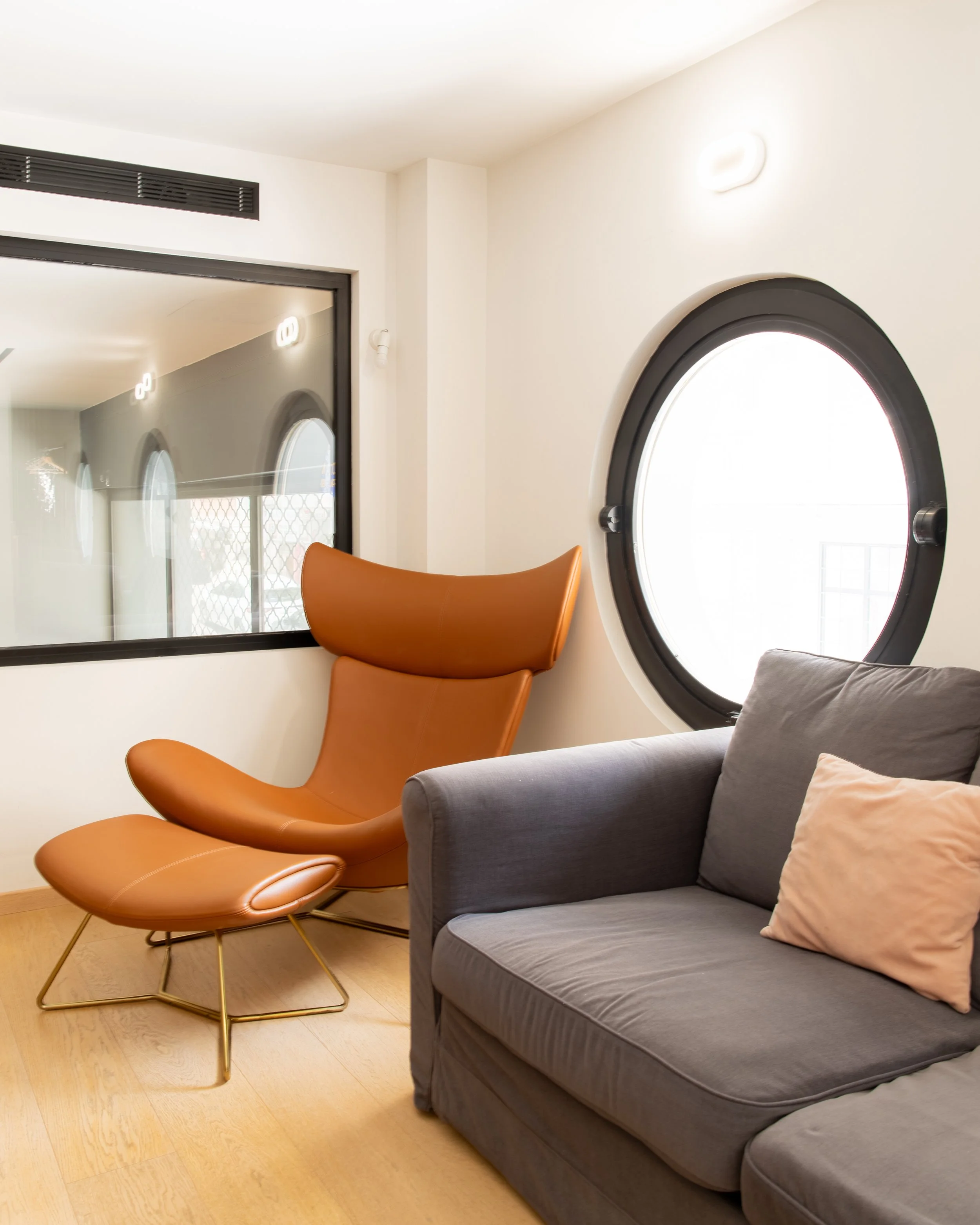 Modern living room with orange lounge chair, gray sofa with pillows, wooden floor, large window with round frame, and glass partition with black frame.