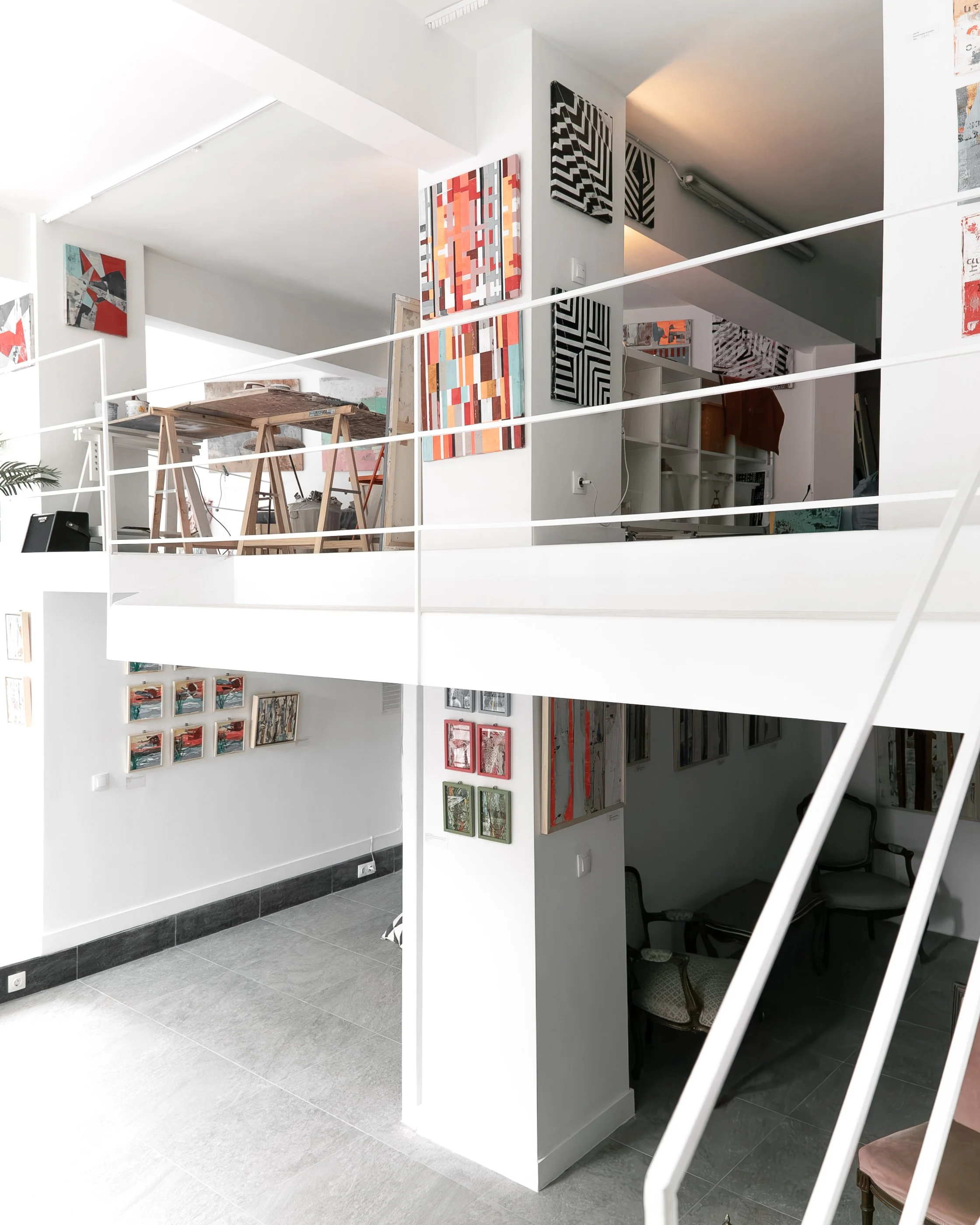 Interior view of an art gallery or studio with paintings on white walls, including abstract geometric art using bright colors and black-and-white patterns, wooden furniture, and a staircase with white railings.