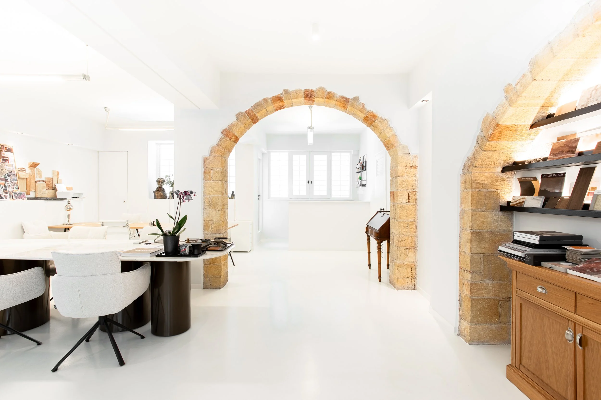 Bright, modern living room with white walls, exposed brick archways, and a mix of contemporary and vintage furniture, including a white dining table with white chairs, a wooden sideboard, and black bookshelves.