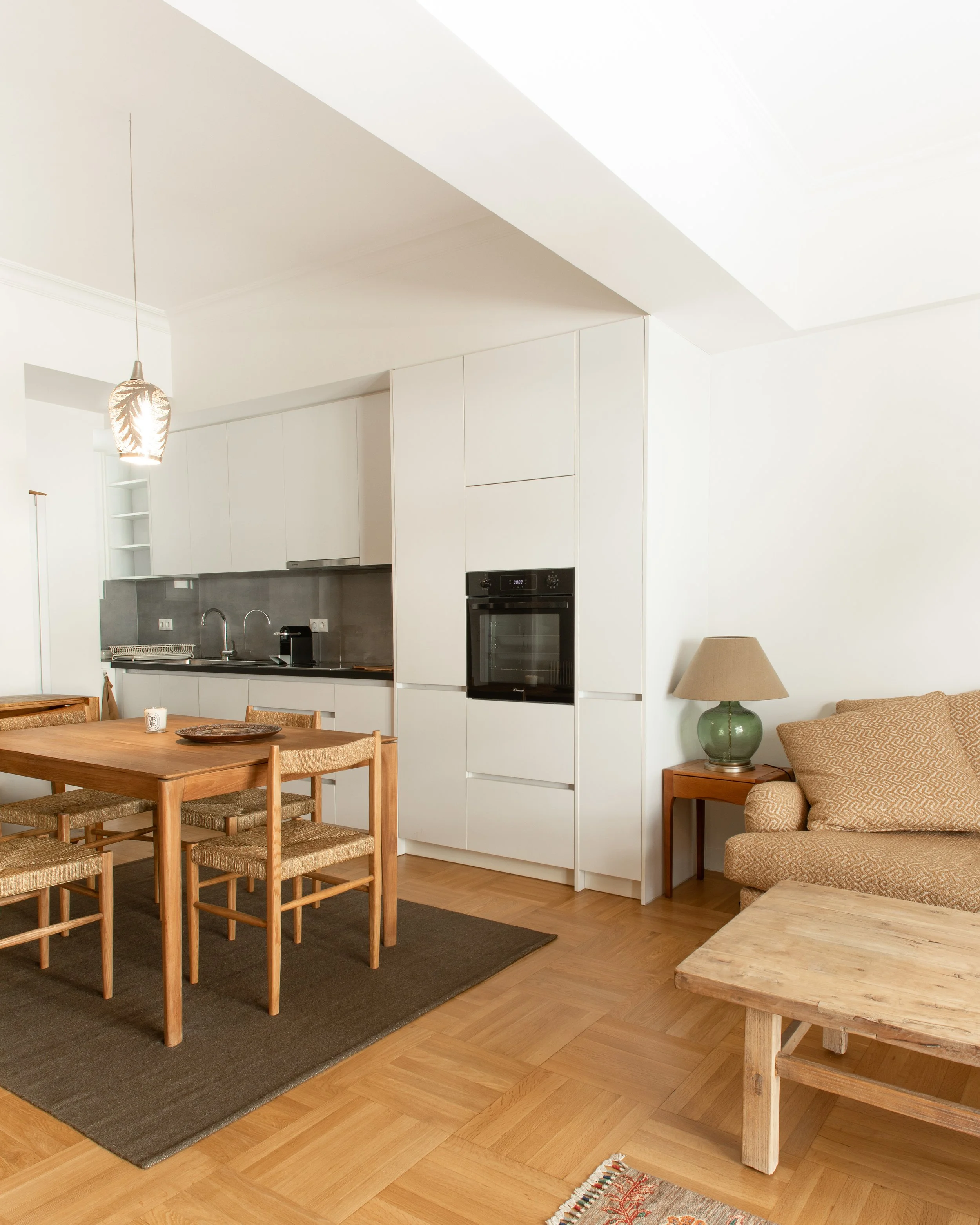 A bright, modern kitchen and dining area with white cabinets, a wooden dining table with six chairs, a black oven, and a pendant light fixture. There is a beige sofa with a side table and lamp nearby, and wooden flooring with a dark rug under the din