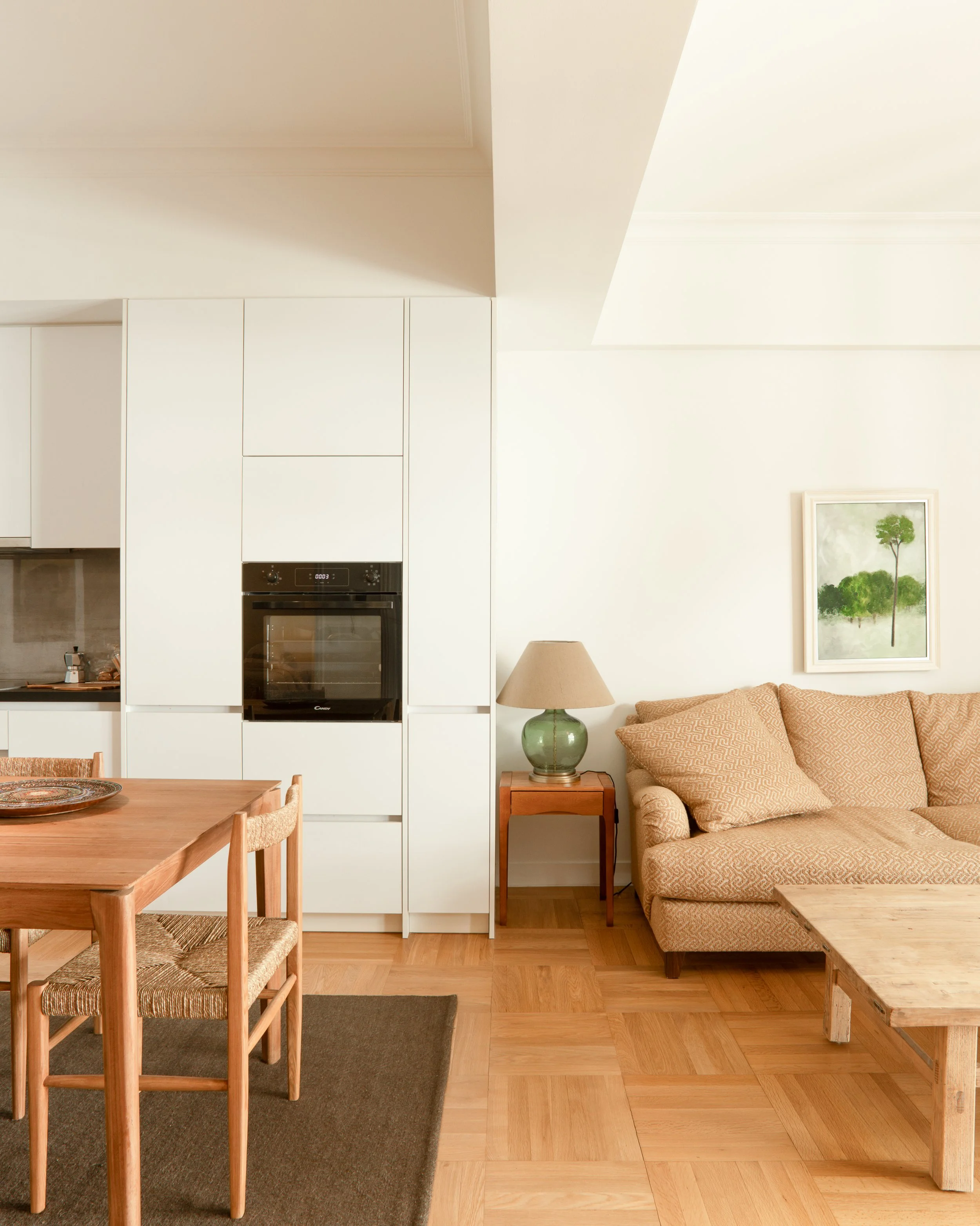 A living room with light wood flooring, a patterned beige sofa, a small wooden side table with a green glass table lamp, a potted plant in a framed print on the wall, a white cabinet and oven, a round wooden dining table with four chairs, and a dark 