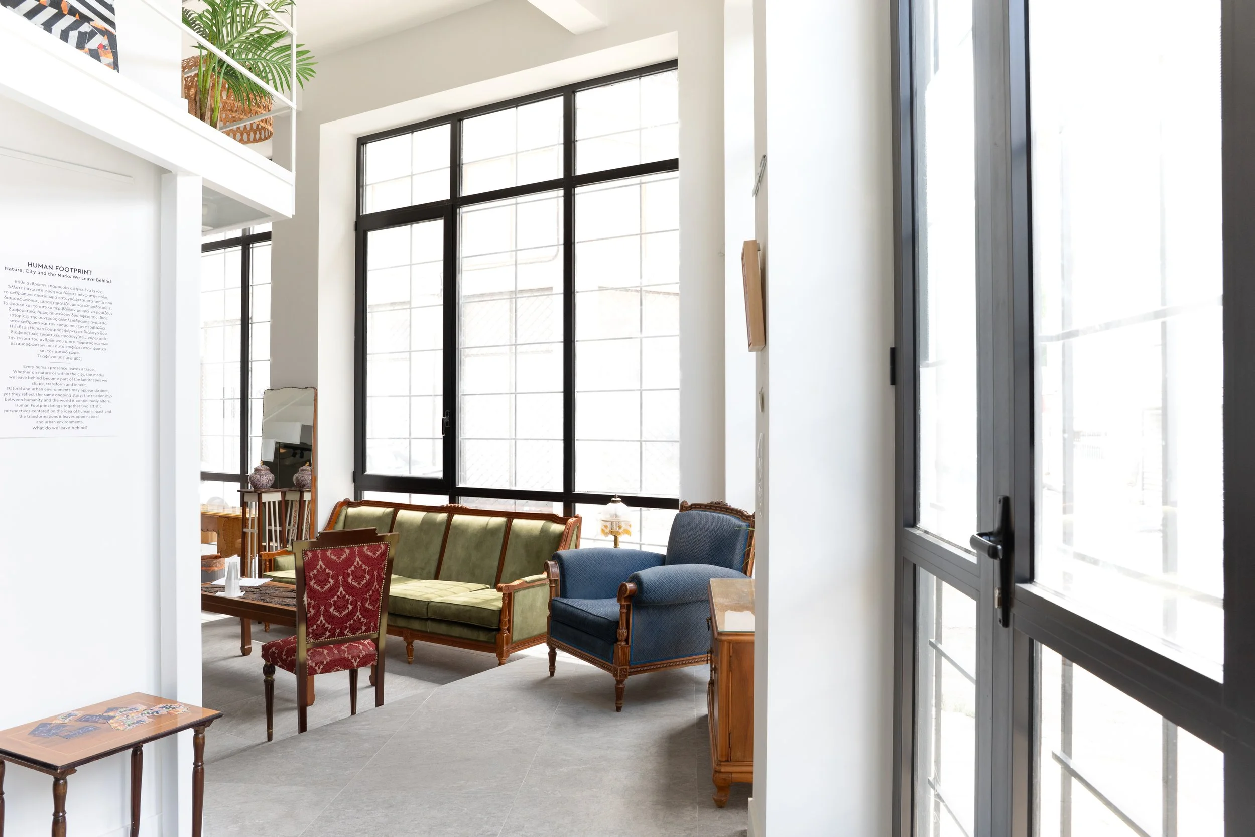 Interior view of a bright, modern living room with large windows, vintage-style furniture including a green velvet sofa, armchairs, and a small table with decorative items. Part of a second-story balcony with a plant is visible.
